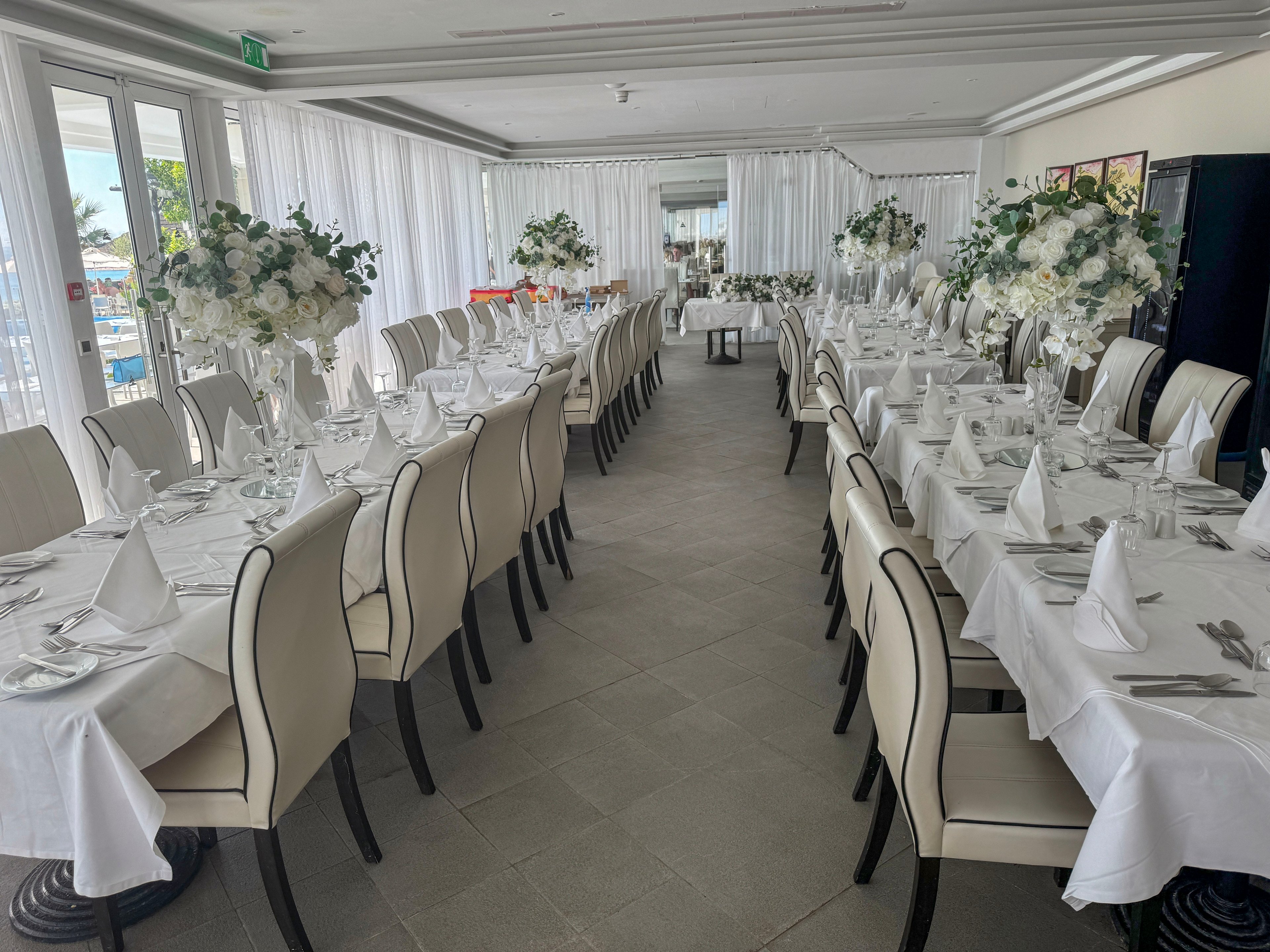 A long, bright banquet hall is set with two rows of white-clothed tables and cream-colored chairs. Each table features neatly folded white napkins, glassware, and cutlery arranged for a formal event. Tall floral centerpieces with white flowers and greenery line the tables, adding a sophisticated touch. Large windows and sheer white curtains create an airy atmosphere, suggesting a waterfront or resort location prepared for a celebration.