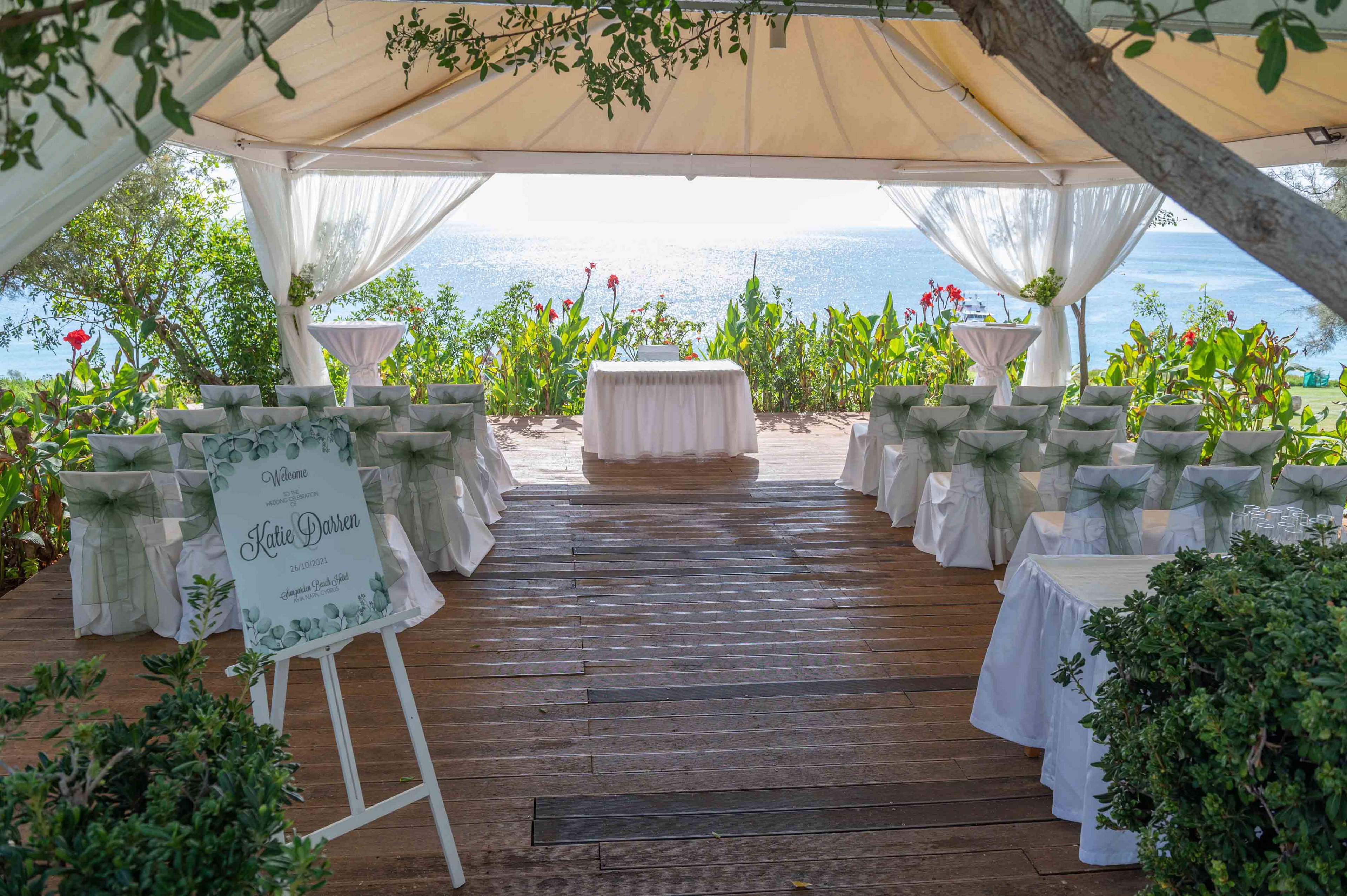 An elegant wedding ceremony area is arranged under a white canopy overlooking the sea. Rows of chairs are decorated with soft green bows, facing a central table at the front. A floral sign on an easel welcomes guests, framed by lush greenery and colorful flowers. Sheer curtains and natural wooden flooring add a romantic, airy atmosphere to the coastal setting.