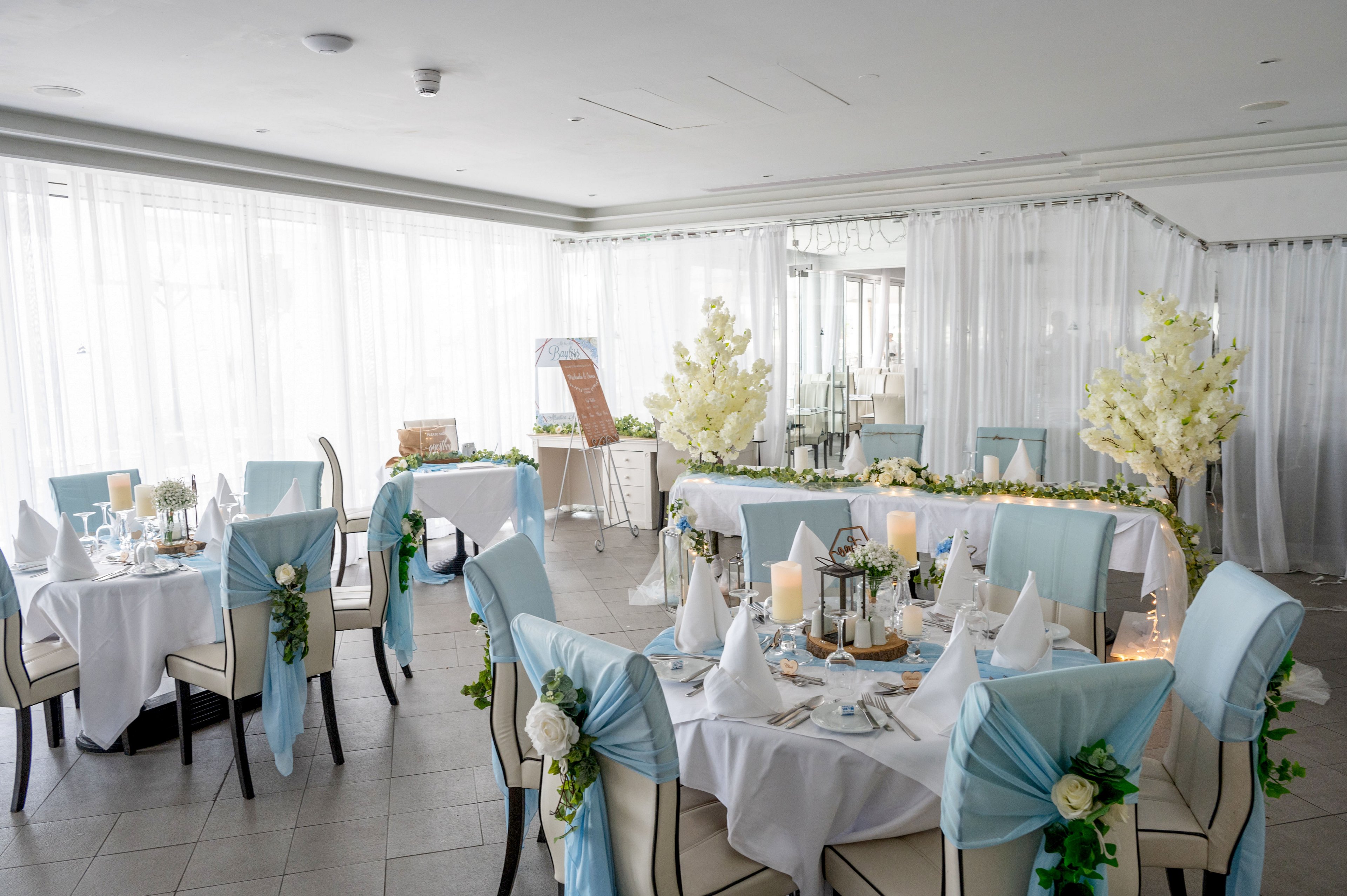 A bright, elegantly decorated reception hall features round tables set for a formal event. White tablecloths and chair covers are accented with soft blue sashes and floral arrangements. Candles, glassware, and folded napkins create a refined, romantic atmosphere. Large windows with sheer curtains and white flower displays enhance the airy, sophisticated setting.