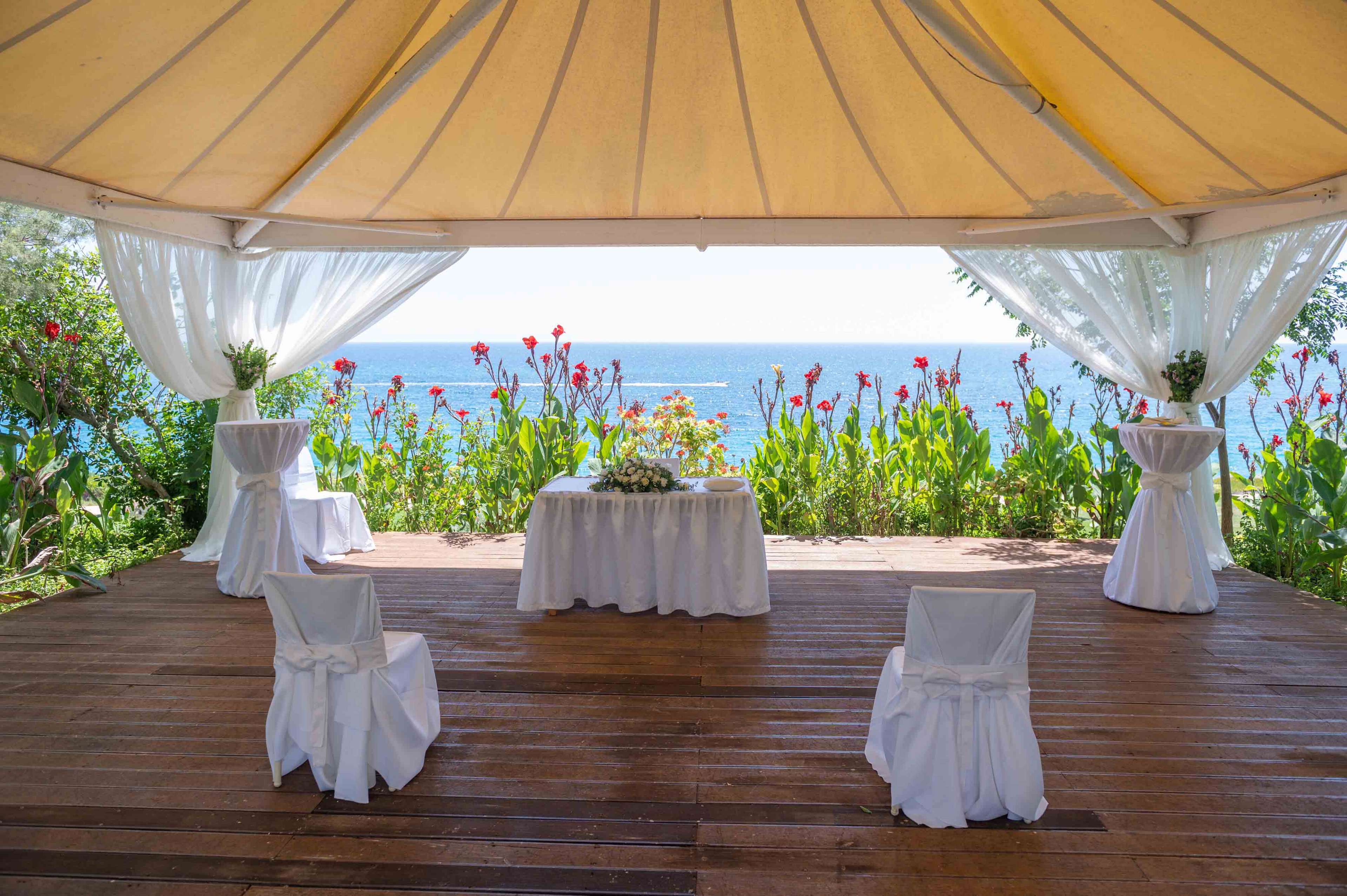 An elegant wedding ceremony setup is arranged under a large gazebo overlooking the sea. White chairs and tables are draped in cloth, with a central table decorated with flowers. Sheer white curtains frame the open sides of the structure, revealing lush green plants and red flowers along the edge. The bright daylight and ocean horizon create a romantic and serene atmosphere.
