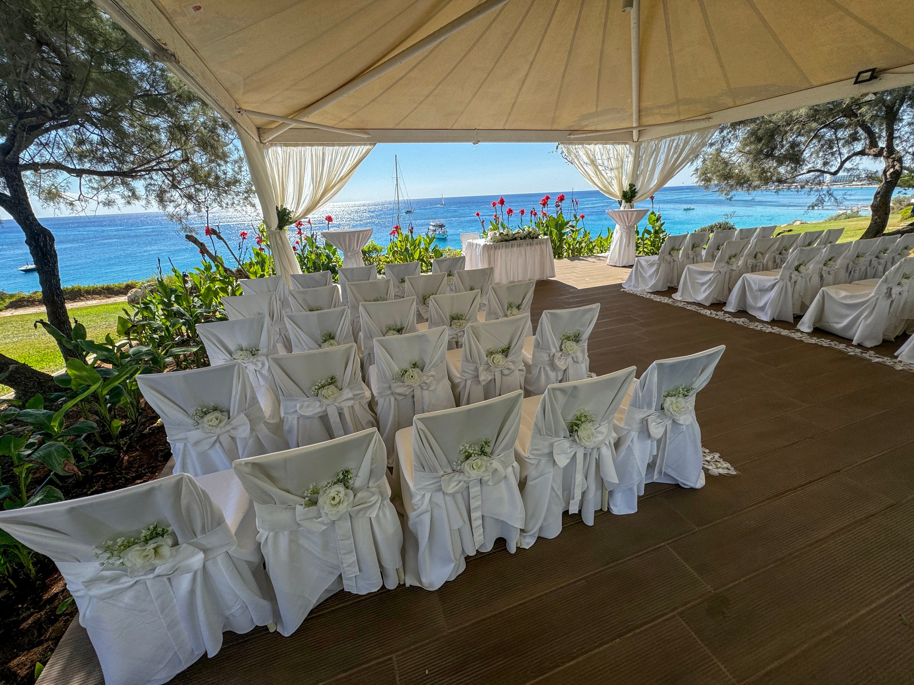 Rows of white-covered chairs are arranged for a wedding ceremony under a large open-sided tent. The venue overlooks a bright blue ocean with sailboats visible in the distance. Green plants and colorful flowers frame the aisle and the altar table at the front. Sunlight filters through the canopy, creating a serene and romantic coastal atmosphere.