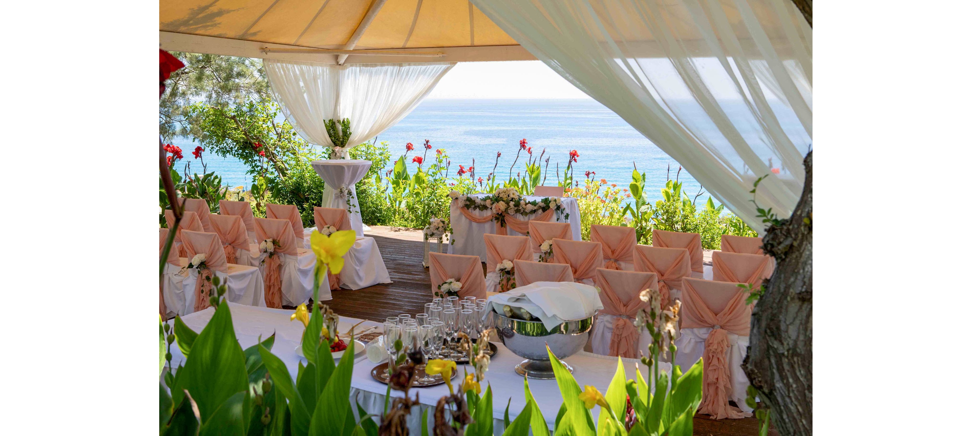 An elegant wedding ceremony area is arranged under a canopy overlooking the sea. Rows of chairs with peach-colored covers and bows face a floral-decorated table. Glassware and serving bowls are prepared on a white-clothed table in the foreground. Lush green plants and bright flowers frame the scene, with calm blue water visible in the background.
