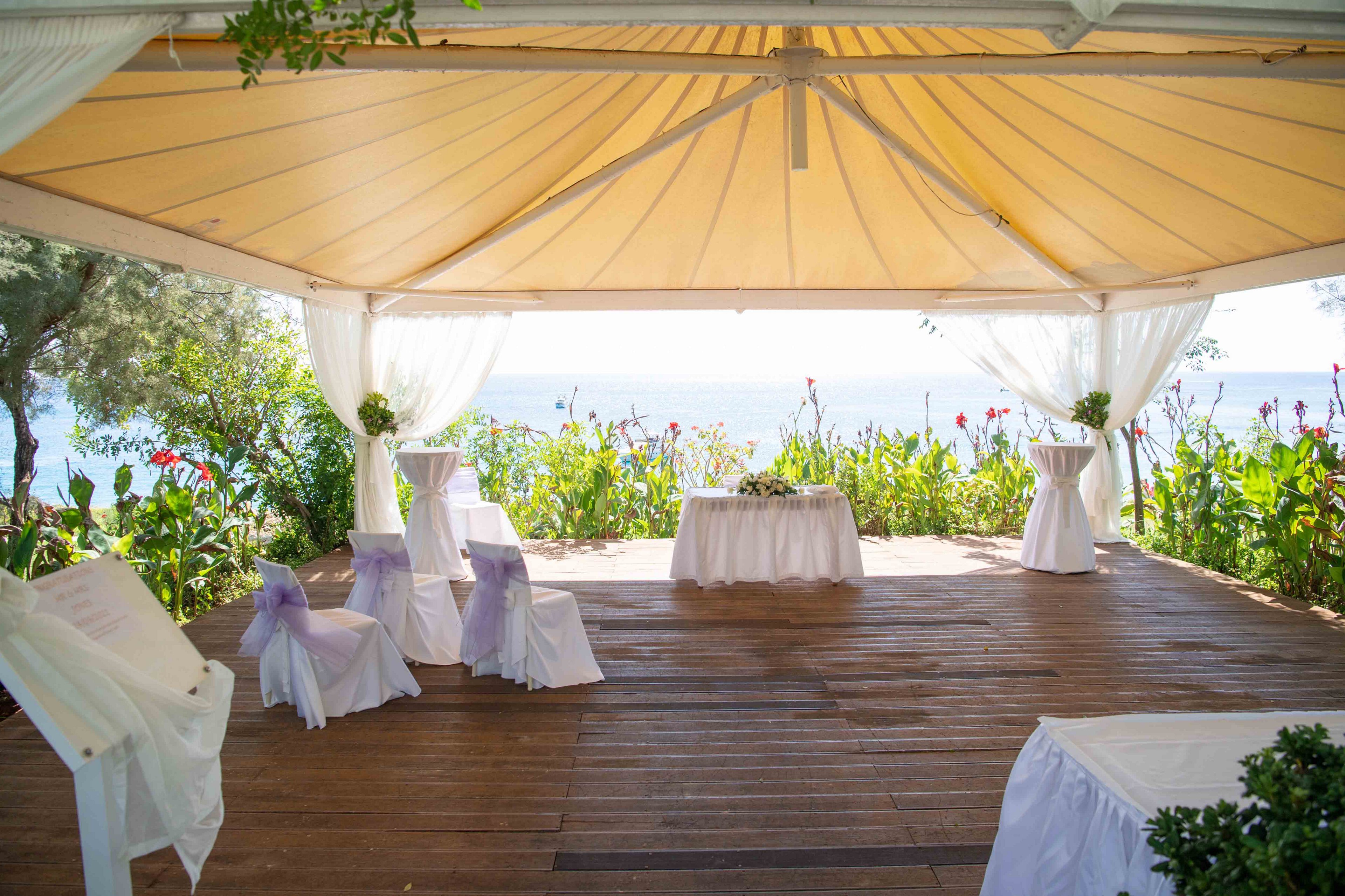 An elegant white gazebo is set up for a wedding ceremony overlooking the sea. White chairs with lavender ribbons face a small table at the front, placed on a wooden deck. Sheer white curtains frame the open sides, revealing lush green plants and colorful flowers around the perimeter. Bright daylight and soft pastel accents create a romantic, airy atmosphere by the water.