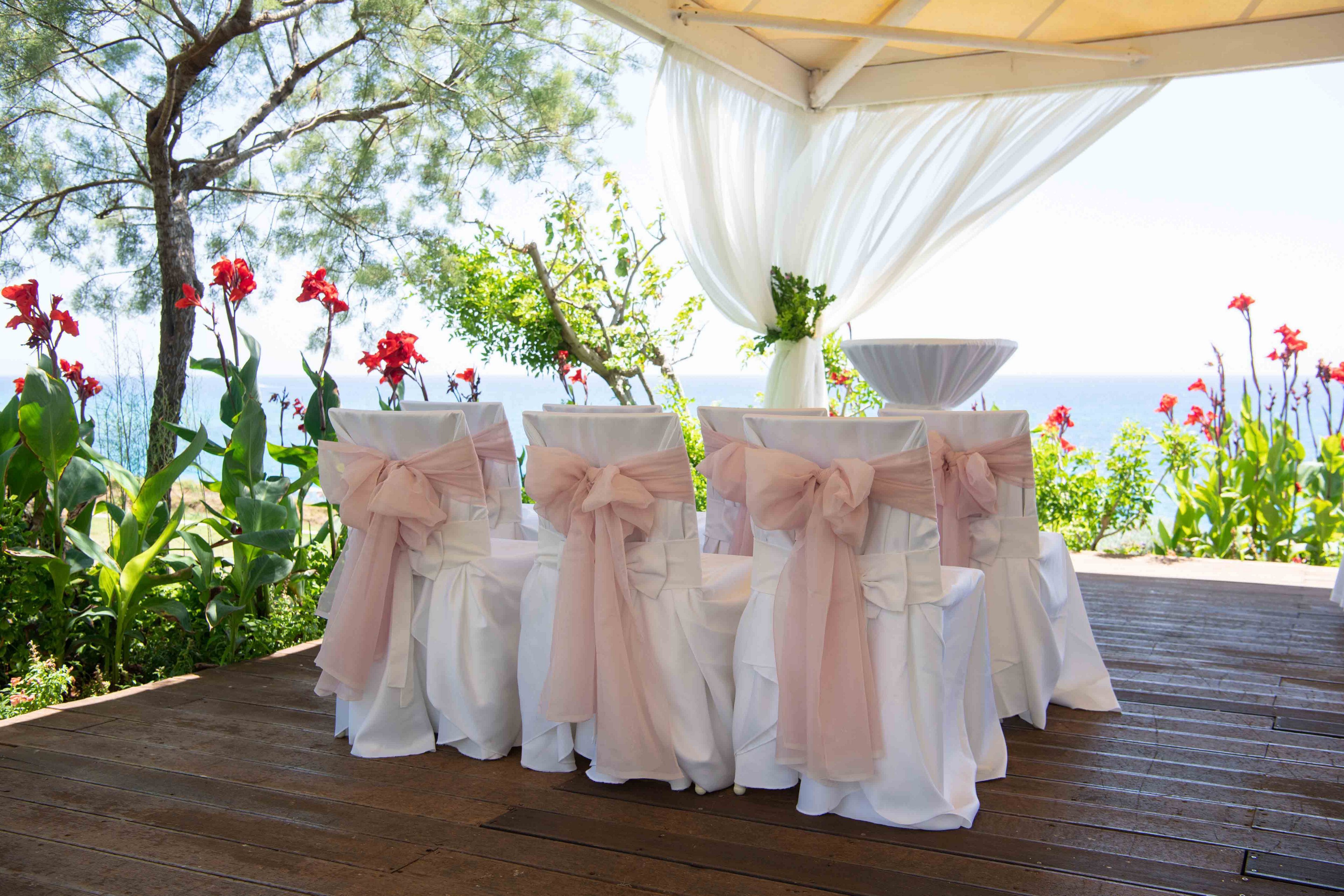 White chairs with soft pink bows are arranged under a canopy for an outdoor wedding ceremony. Sheer white drapes frame the view, gently flowing in the breeze. Bright red flowers and lush green plants surround the wooden deck, with the ocean visible in the background. The scene feels elegant, romantic, and summery.
