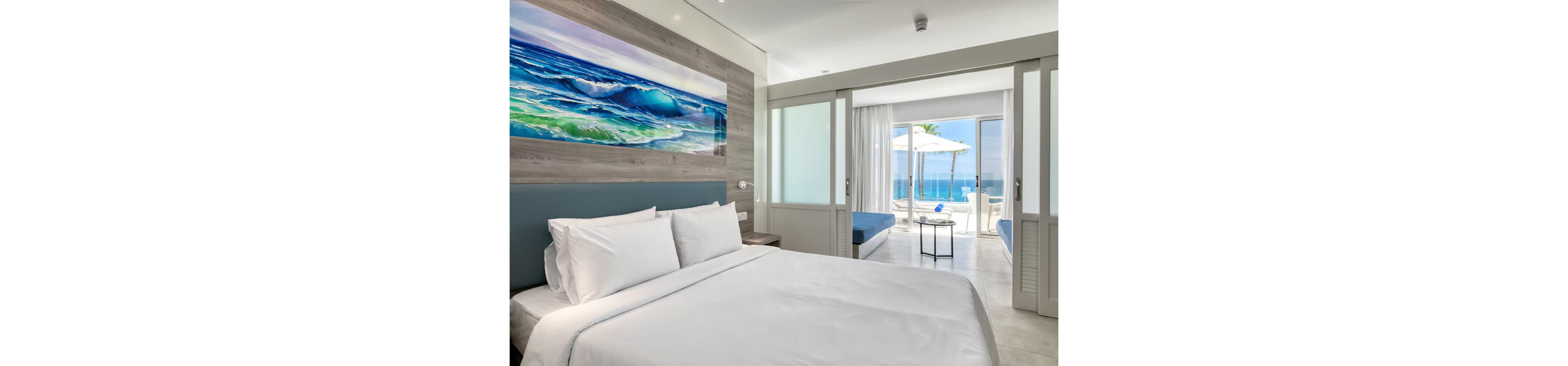 Modern hotel room with a neatly made bed faces sliding glass doors that open to a terrace overlooking the ocean. A large colorful painting of waves hangs above the headboard, echoing the seaside setting. The space features light neutral tones, minimalist furniture, and abundant natural light. Outside, lounge chairs and a sun umbrella create a relaxing beachfront atmosphere.