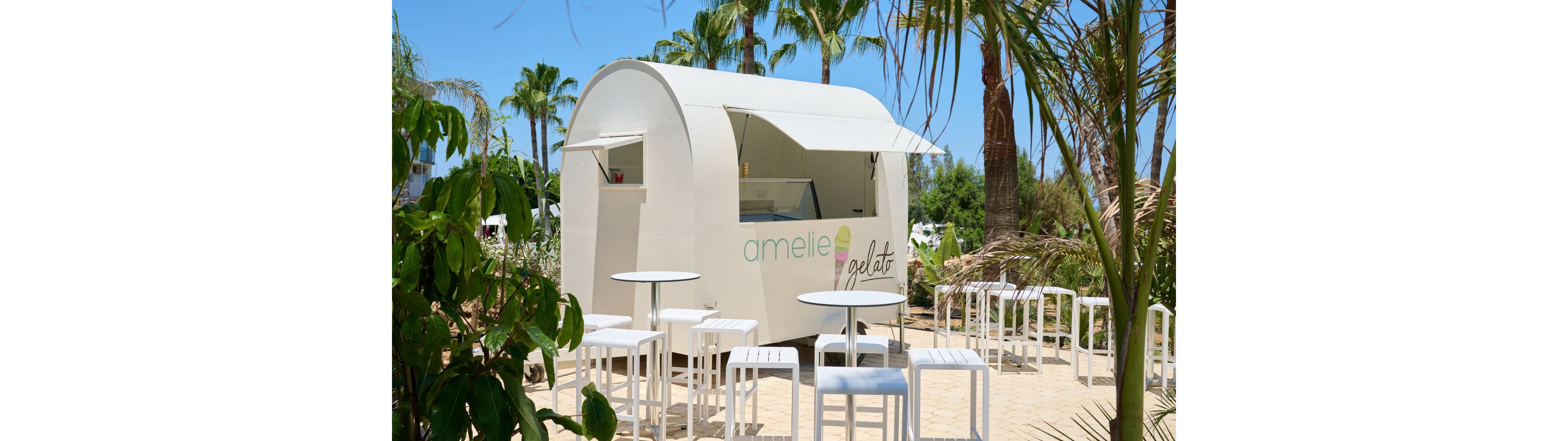 A sleek white gelato kiosk stands outdoors surrounded by palm trees and tropical plants. Several white tables and stools are arranged on a sandy patio in front of the stand, creating a casual seating area. The sky is clear and blue, suggesting a warm, sunny day. The scene feels relaxed and inviting, ideal for enjoying cold treats in a resort-like environment.