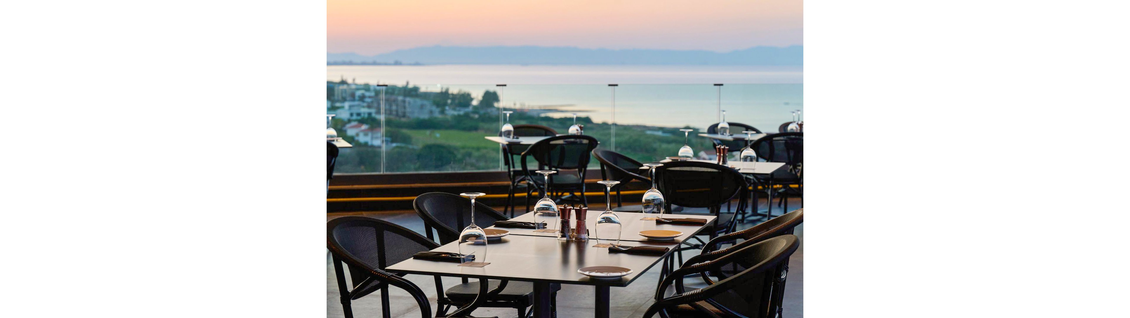 An elegant rooftop restaurant terrace is set with dark tables and chairs overlooking a calm sea at sunset. Wine glasses are placed upside down on the tables alongside plates, cutlery, and condiments. The glass railing frames a scenic coastal town and distant hills under a pastel sky. The atmosphere feels quiet, refined, and ready for evening diners.