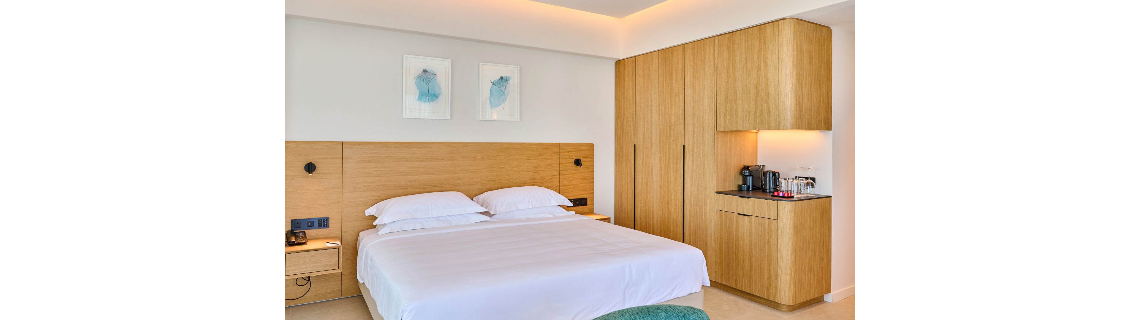 A contemporary hotel bedroom features a large bed with crisp white linens and double pillows. The room is designed with light wood paneling that extends from the headboard to a built-in wardrobe. A compact coffee station with a coffee machine and cups is set into a corner cabinet. Soft indirect ceiling lighting and two framed artworks above the bed create a calm, minimalist atmosphere.
