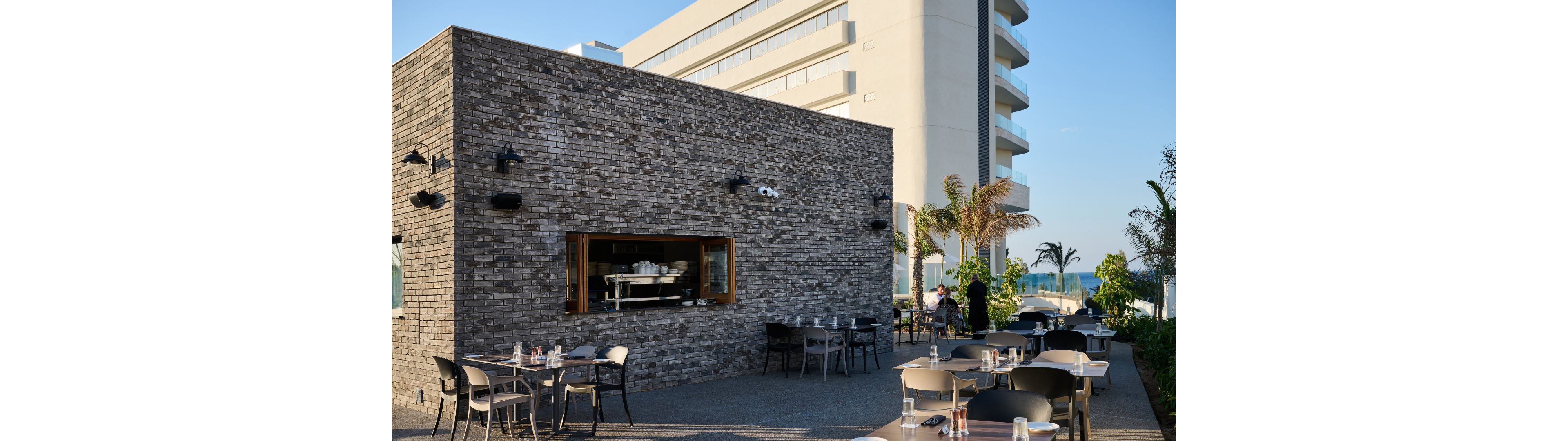 An outdoor restaurant terrace features neatly arranged tables and chairs beside a modern brick building. Glassware and place settings are prepared on the tables, suggesting readiness for guests. In the background, a contemporary multi-story hotel and palm trees indicate a coastal or resort setting. Warm sunlight and clear skies create a relaxed, inviting atmosphere for al fresco dining.
