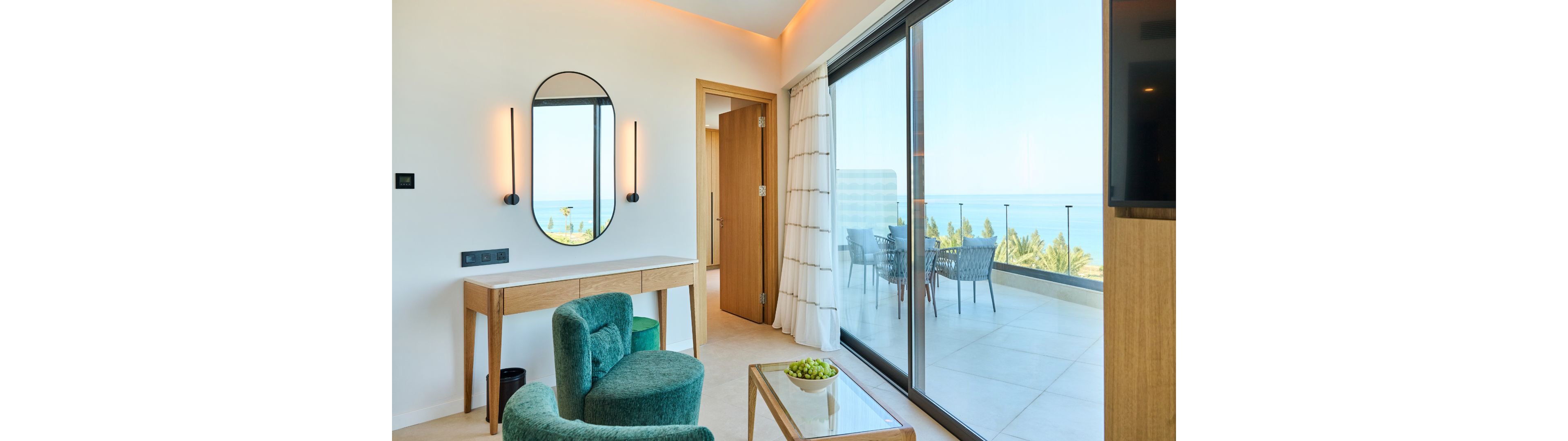 A contemporary hotel room features a teal armchair and a wooden vanity with a round mirror and wall lights. Large sliding glass doors open onto a spacious balcony furnished with a table and chairs. Beyond the balcony, the ocean and palm trees are visible under a clear sky. Soft neutral tones and natural light create a calm, relaxing atmosphere.
