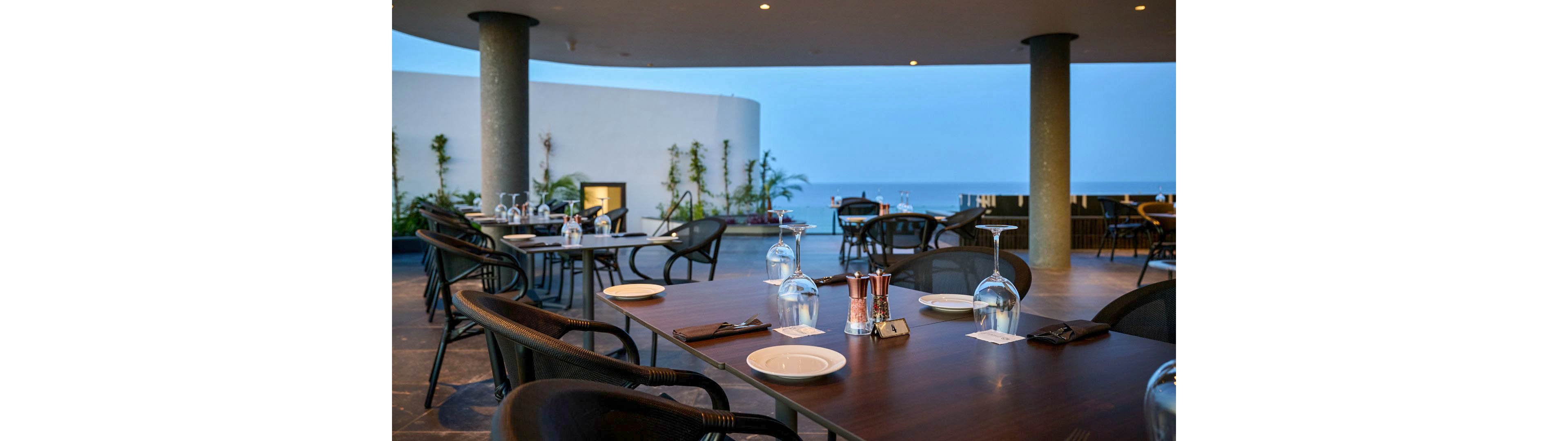 An open-air restaurant terrace is set with dark wooden tables and neatly arranged glassware. Empty chairs surround the tables, suggesting a calm moment before guests arrive. Large columns frame the view of the ocean and a soft blue evening sky in the background. Subtle lighting and minimalist architecture create a relaxed, upscale dining atmosphere.