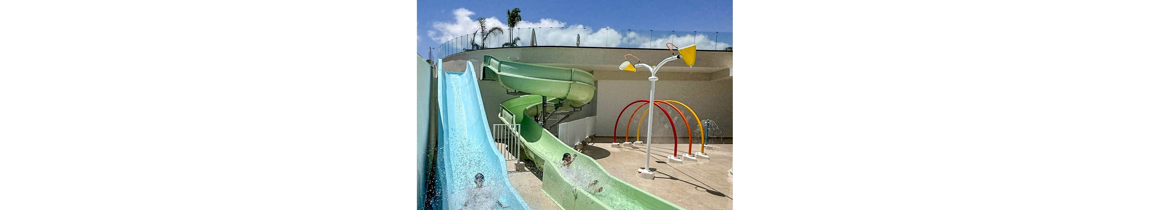 Two children slide down parallel water slides at an outdoor water park. One slide is straight and light blue, while the other is a winding green flume. In the background, colorful rainbow-shaped spray arches and a tall water feature add to the playful scene. The sky is clear and blue, suggesting a sunny, warm day perfect for water activities.