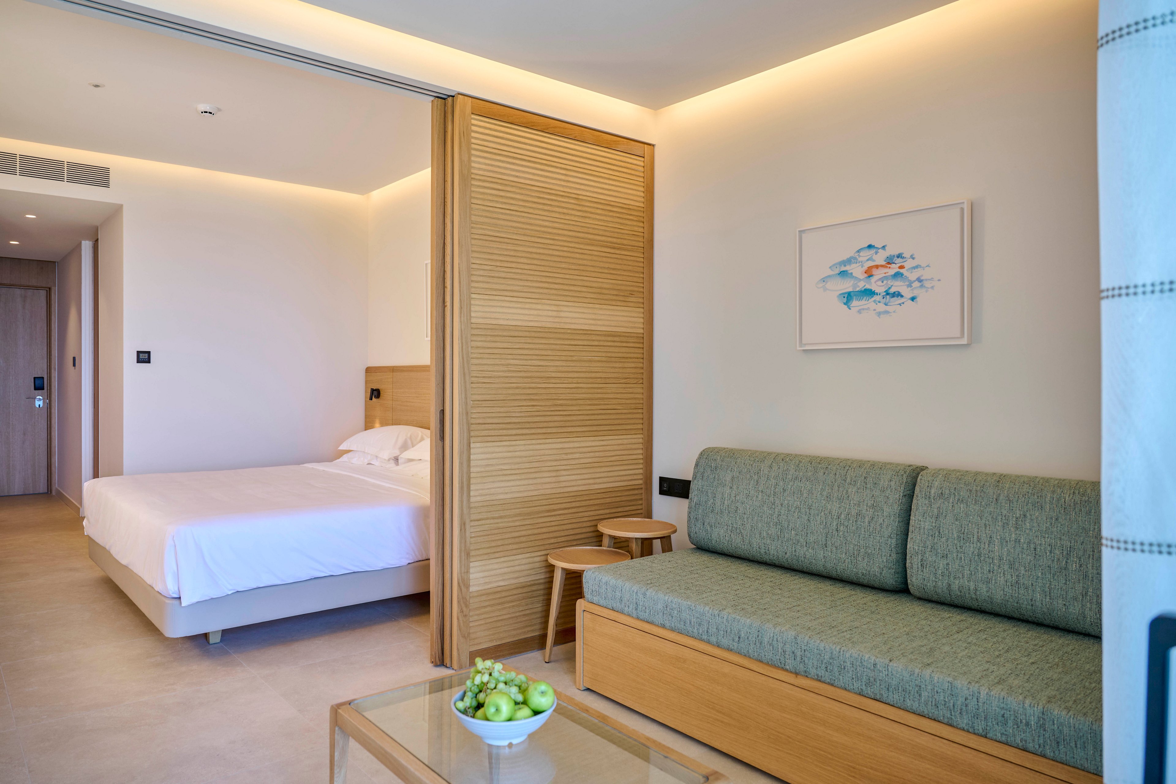 Contemporary hotel room interior featuring a neatly made double bed and a green upholstered sofa. A wooden sliding partition separates the sleeping area from the small lounge space. Soft indirect lighting and neutral walls create a calm, minimalist atmosphere. A glass coffee table with a plate of green grapes adds a fresh accent to the clean design.