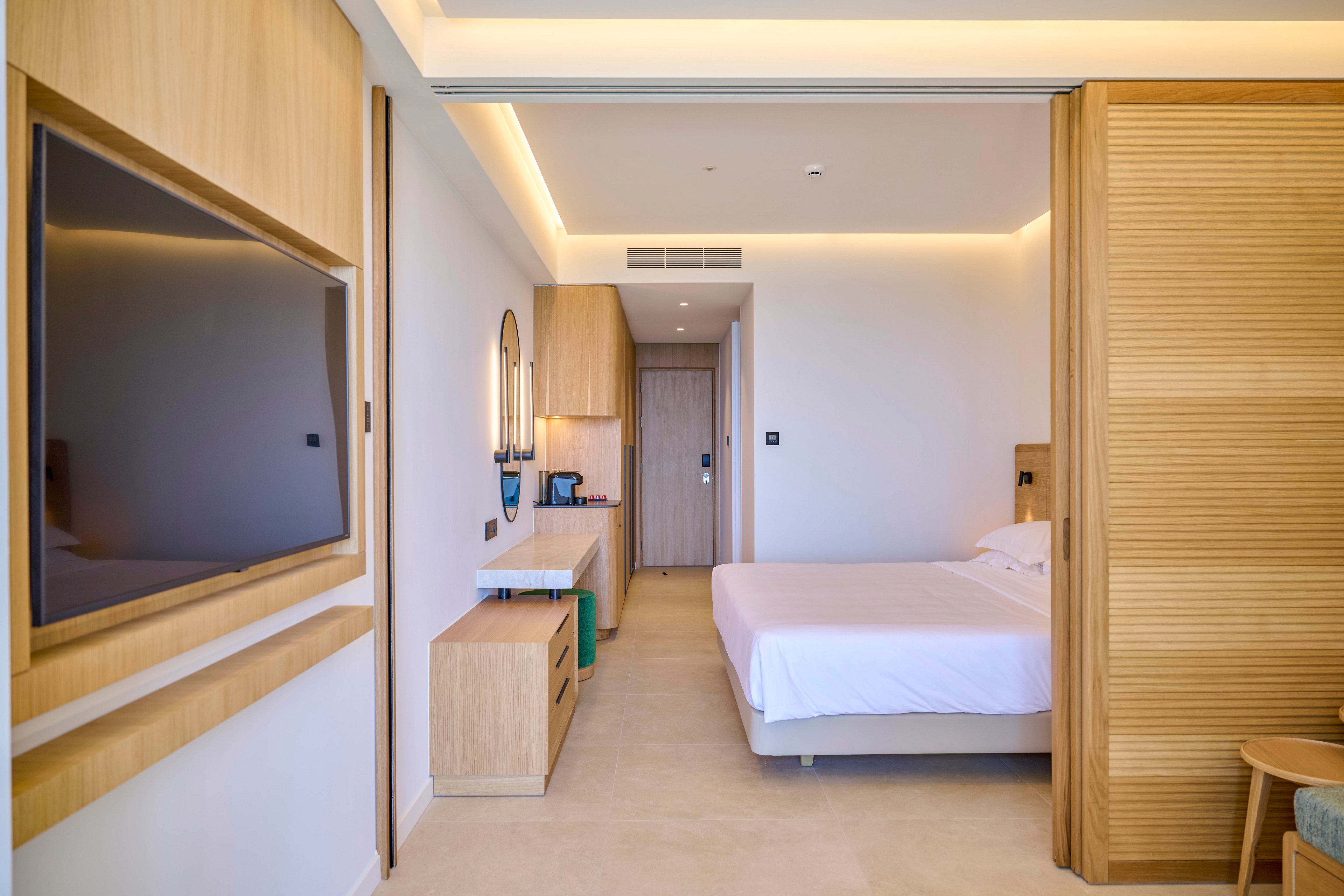 Contemporary hotel room interior featuring a neatly made double bed and light wood furnishings. A large flat-screen television is mounted on the wall opposite the bed. Built-in cabinetry with a desk, mirror, and coffee station extends toward the entrance door. Soft recessed ceiling lighting and neutral tones create a calm, minimalist atmosphere. Sliding wooden panels partially separate the sleeping area from the rest of the room.