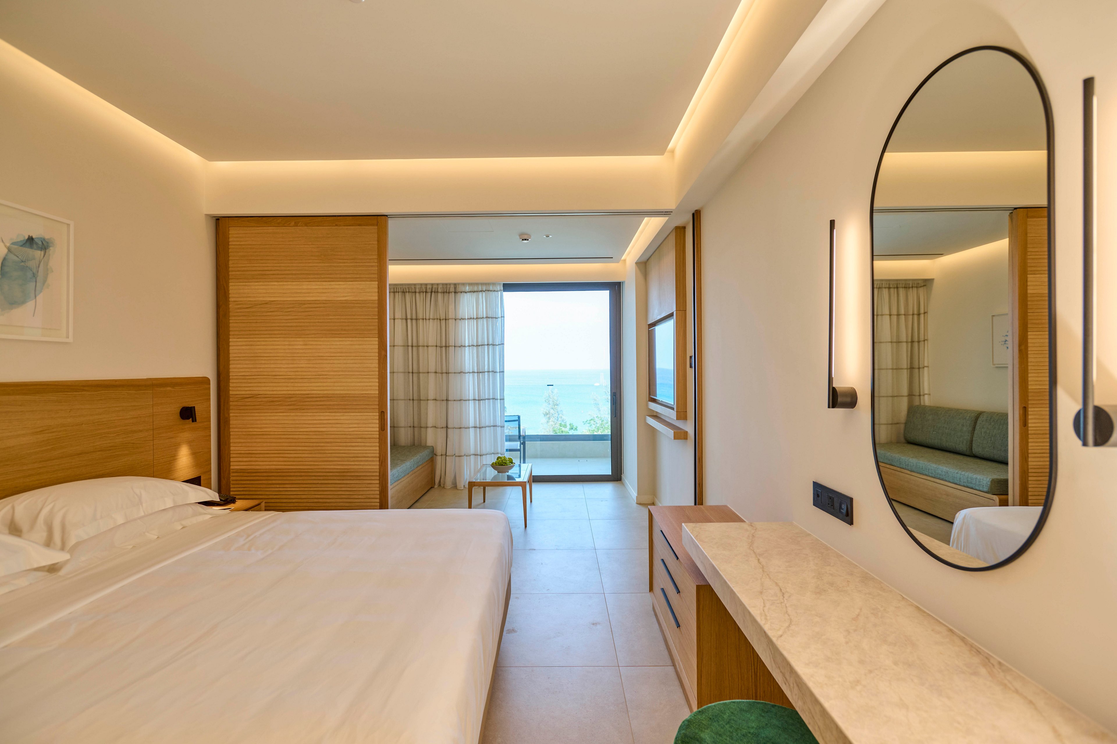 A contemporary hotel bedroom features a neatly made bed with white linens and wooden headboard. The room opens toward a balcony with a view of the ocean framed by large glass doors. Built-in wooden furniture, including a desk and sliding panels, adds warmth to the minimalist design. Soft recessed lighting and a round wall mirror create a calm, inviting atmosphere.