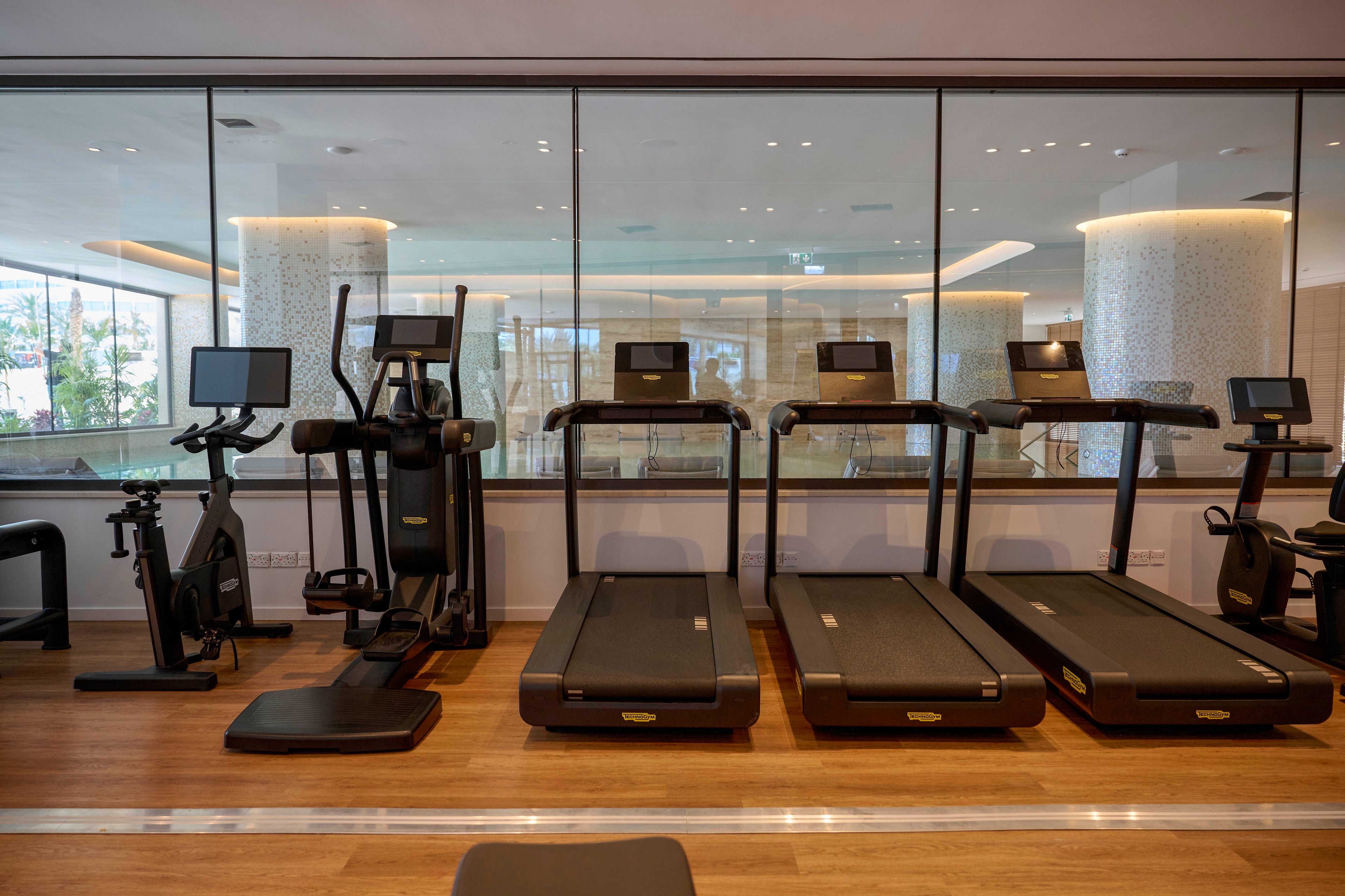 A row of treadmills and cardio machines is arranged neatly in a modern indoor gym. Large floor-to-ceiling windows reveal a bright, upscale interior beyond the glass partition. The wooden flooring and warm lighting create a clean, inviting workout environment. No people are present, emphasizing the equipment and spacious layout.