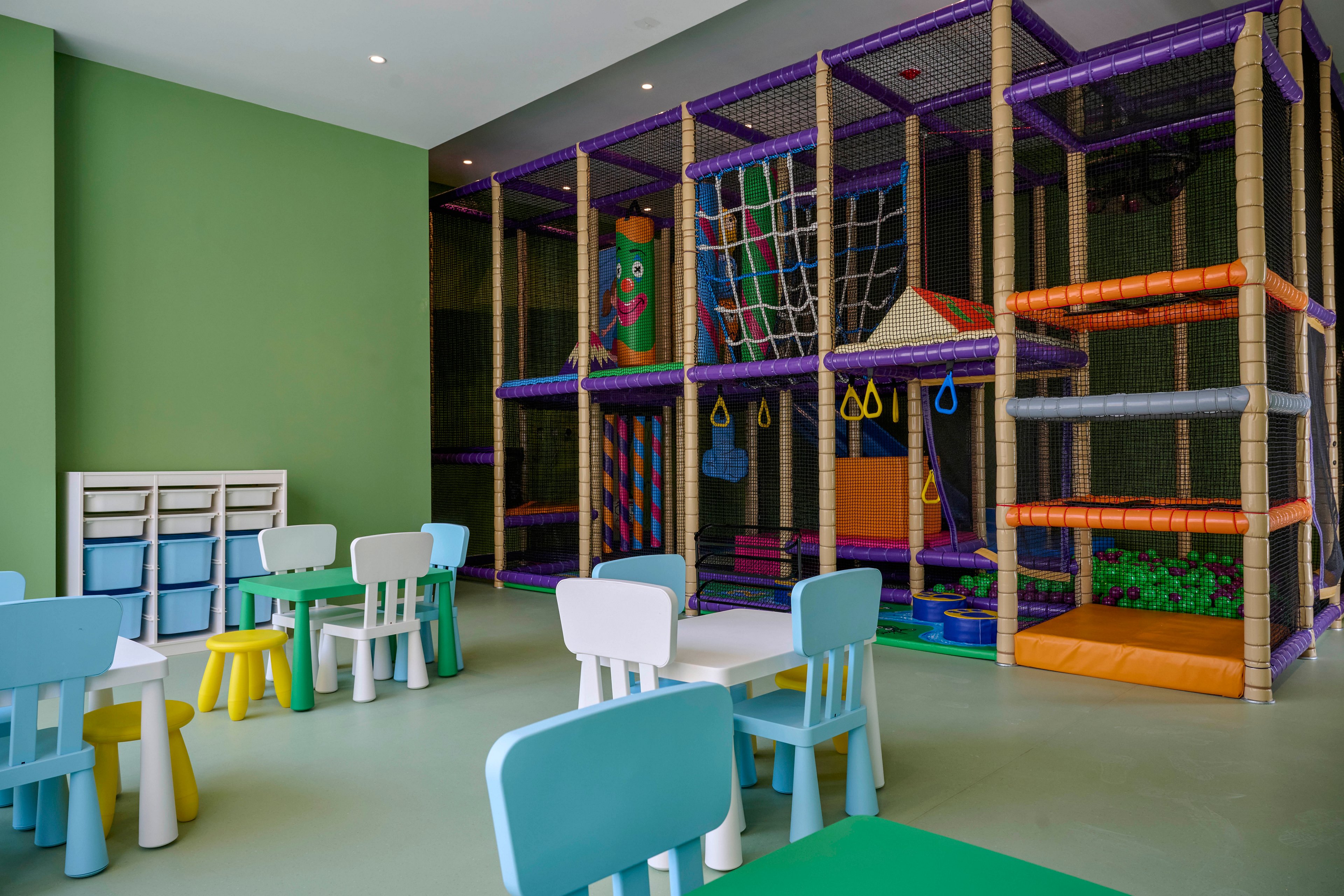 A bright indoor playroom features a large multi-level climbing structure with nets, slides, and soft obstacles. In the foreground, small colorful tables and chairs are arranged for children’s activities. The walls are painted in soft green tones, and storage cubbies sit along one side of the room. The overall scene feels safe, playful, and designed for young children’s recreation and learning.