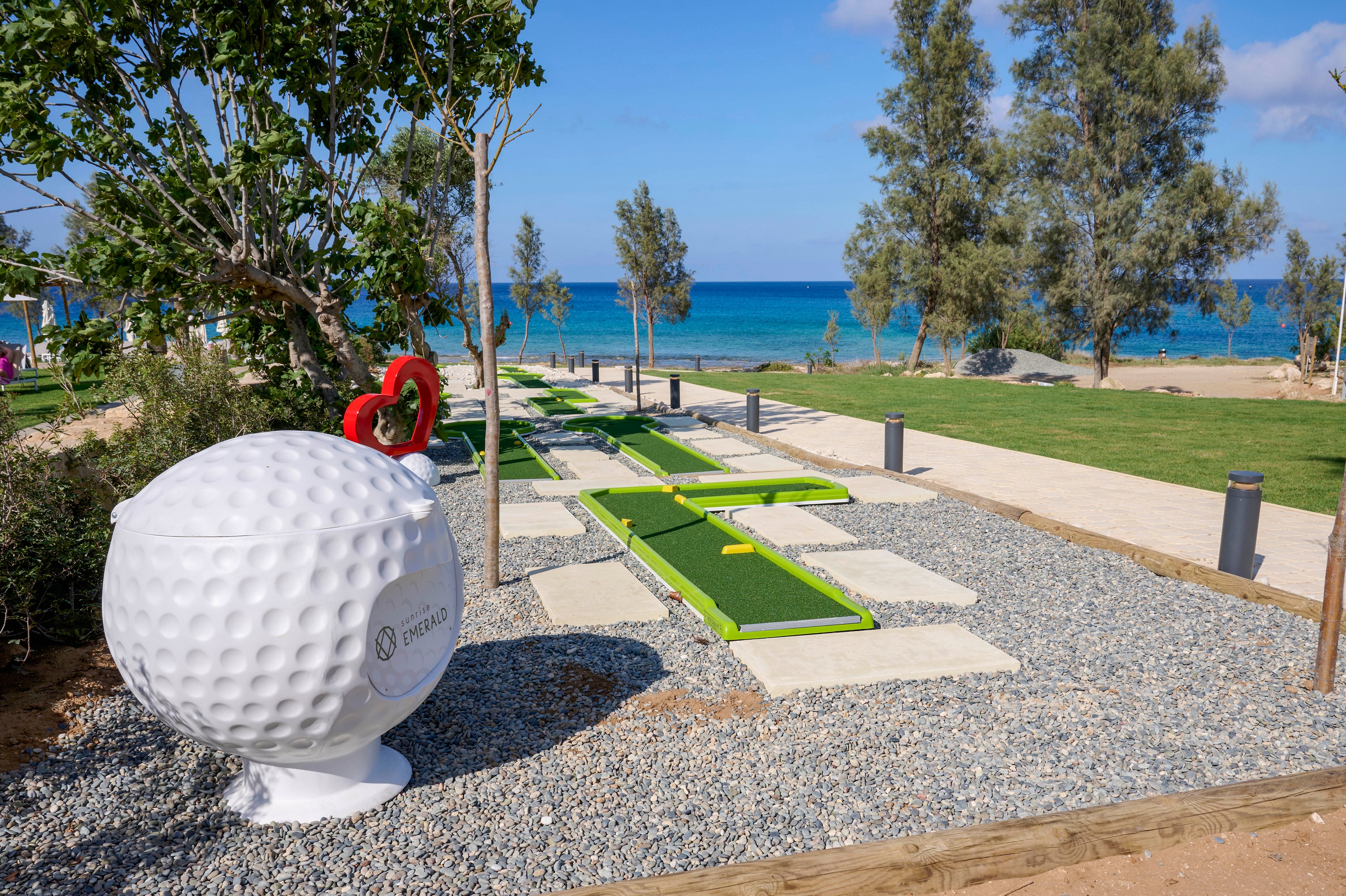 A seaside mini golf course is set against a bright blue ocean backdrop. In the foreground, a large decorative golf ball sculpture with a red heart accent stands beside the pathway. The course features narrow green putting lanes bordered by white stepping stones and gravel. Tall trees, manicured grass, and clear skies create a relaxed holiday atmosphere by the water.