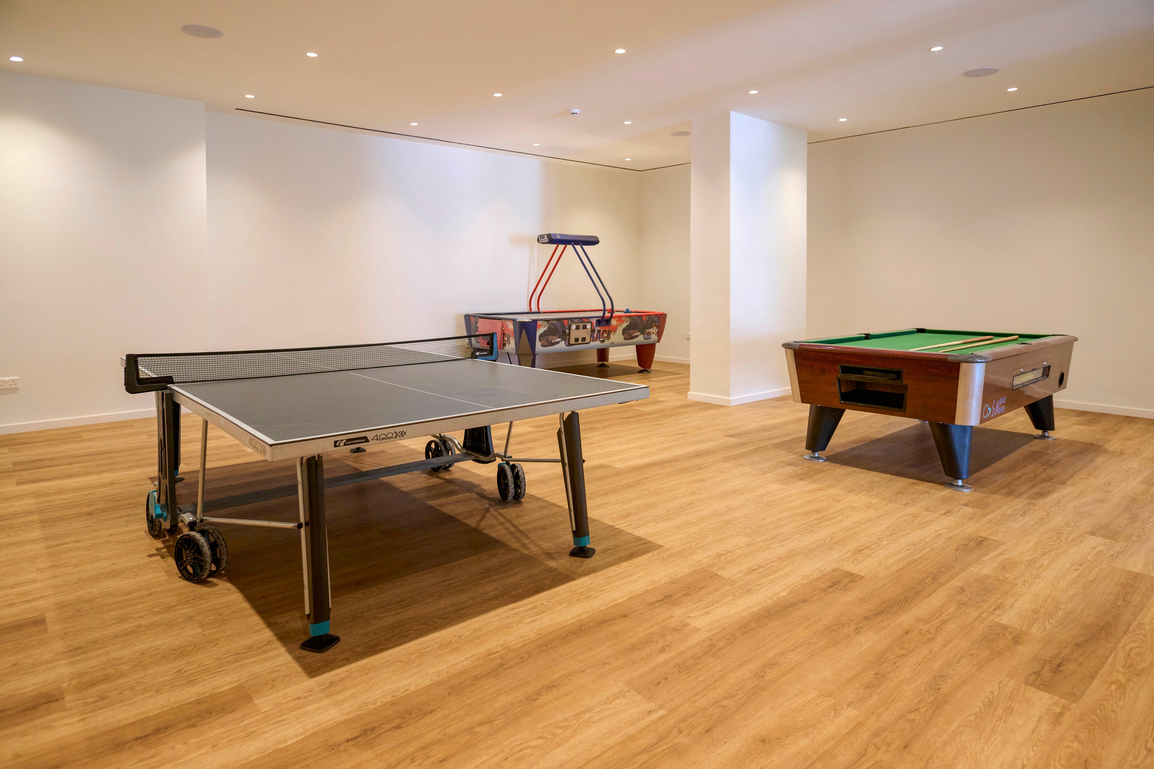 A spacious modern game room features a table tennis table in the foreground. In the background, an air hockey table and a pool table are arranged along the walls. The room has light-colored wooden flooring, white walls, and recessed ceiling lights that create a clean, minimalist look. The space appears uncluttered and designed for indoor recreation and leisure.