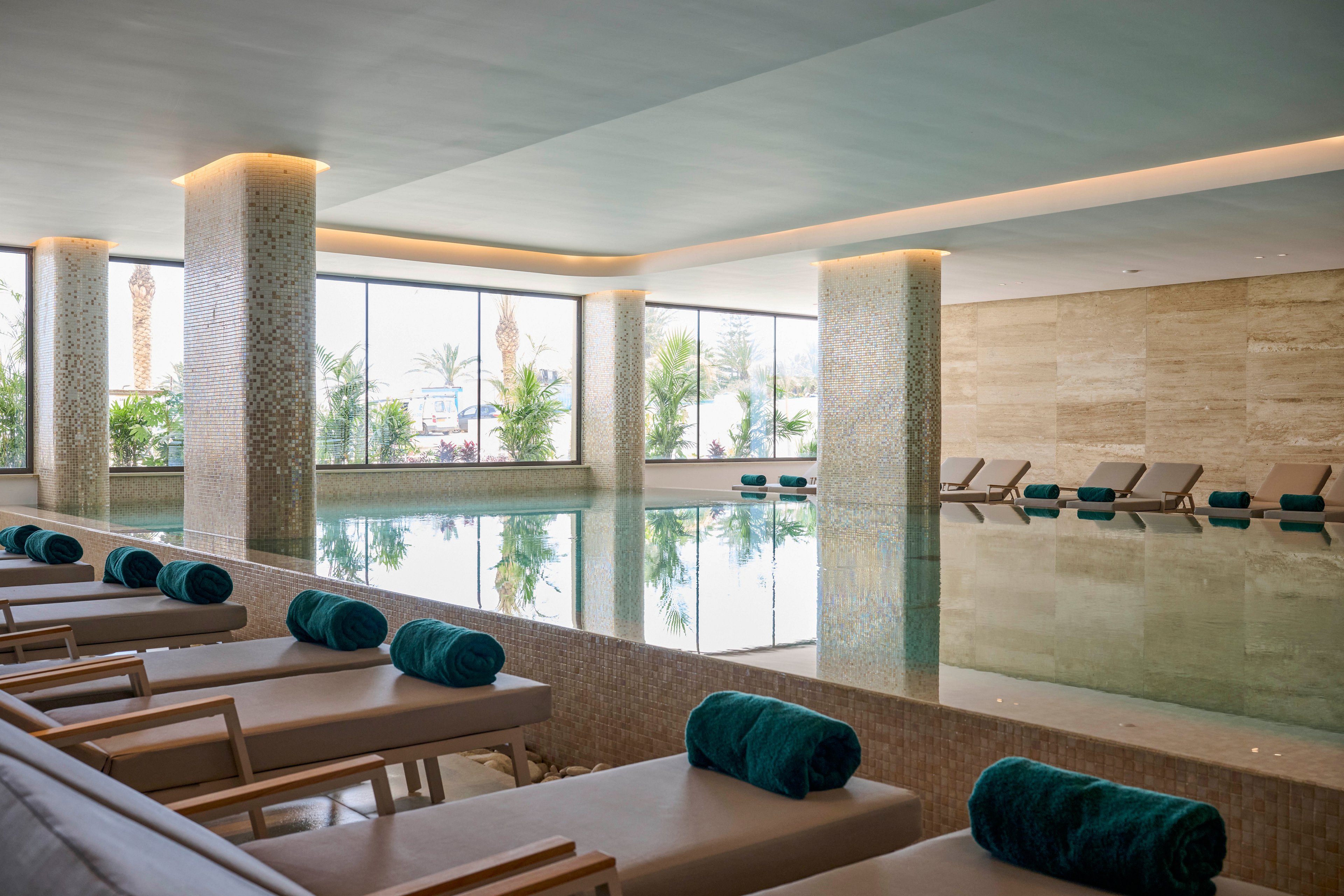 A serene indoor spa pool is surrounded by cushioned lounge chairs with neatly rolled teal towels. Large floor-to-ceiling windows let in natural light and reveal lush greenery outside. Stone columns and neutral-toned walls create a minimalist, upscale atmosphere. The calm water and soft lighting emphasize relaxation and wellness in a resort-like setting.