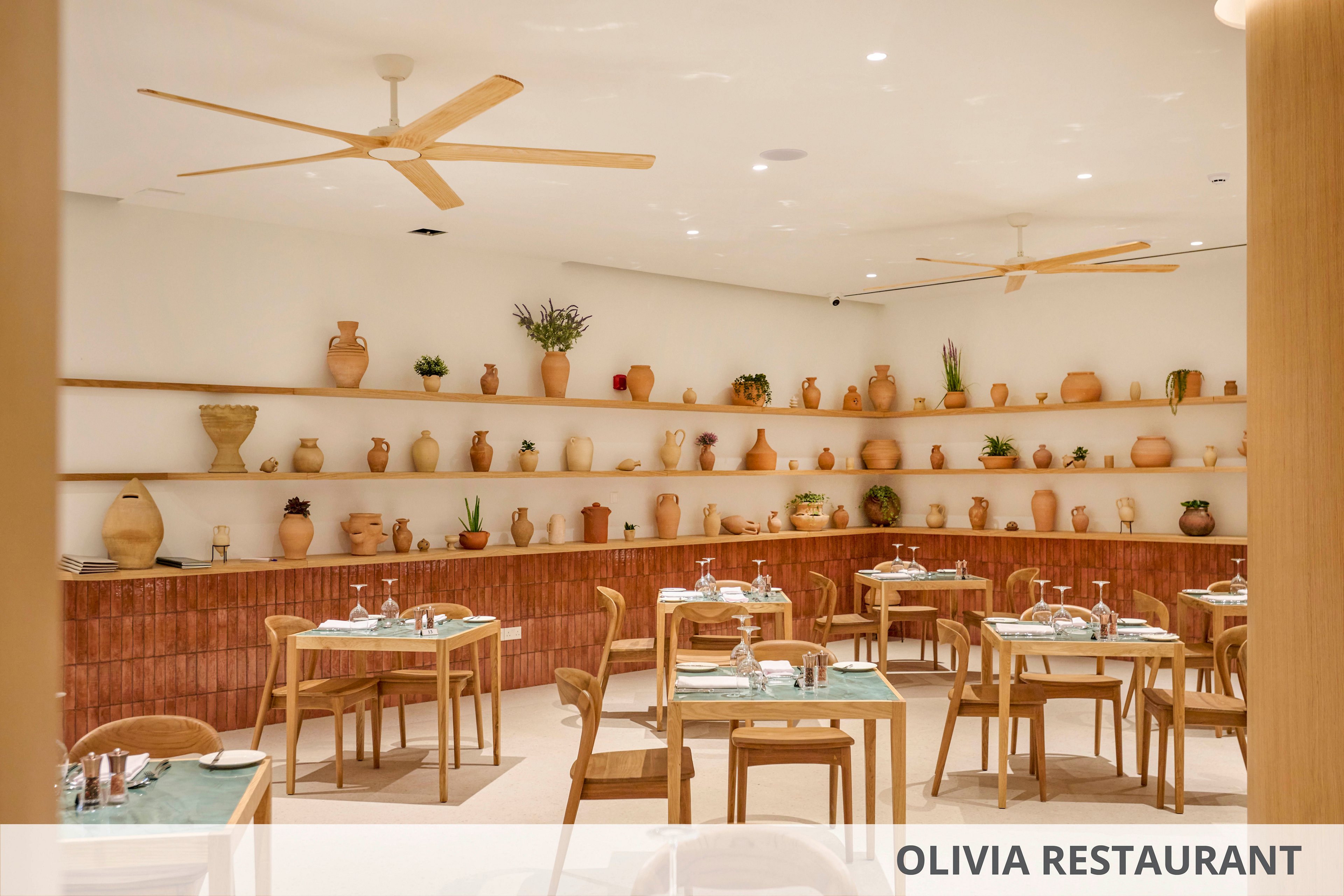 A contemporary restaurant dining room features neatly arranged wooden tables and chairs set for guests. The back wall is lined with long shelves displaying numerous terracotta vases, pots, and decorative objects. Soft neutral tones, warm wood, and green tabletops create a calm, minimalist atmosphere. Ceiling fans and subtle lighting contribute to the relaxed, modern dining setting.