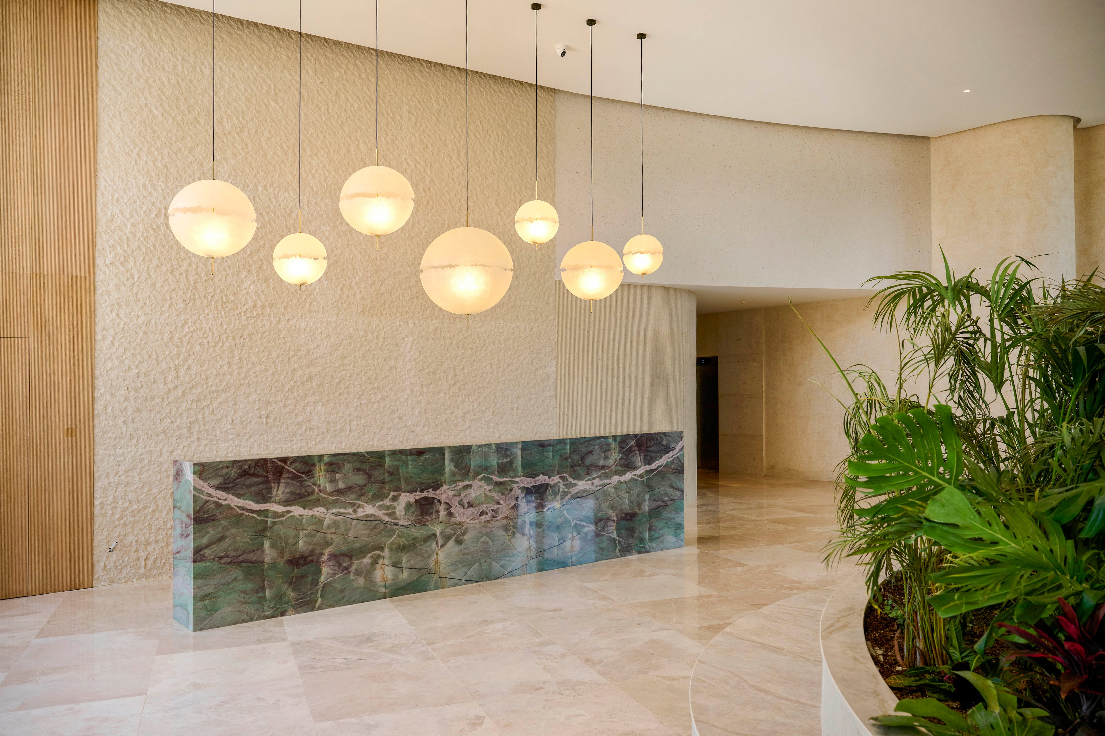 A contemporary lobby interior features a sleek marble reception desk with green and white veining. Several spherical pendant lights hang in a row above the counter, casting a warm glow on the textured stone walls and polished floor. The space is minimalistic and open, with neutral tones and curved architectural lines. A cluster of indoor plants adds a touch of greenery near the entrance corridor.