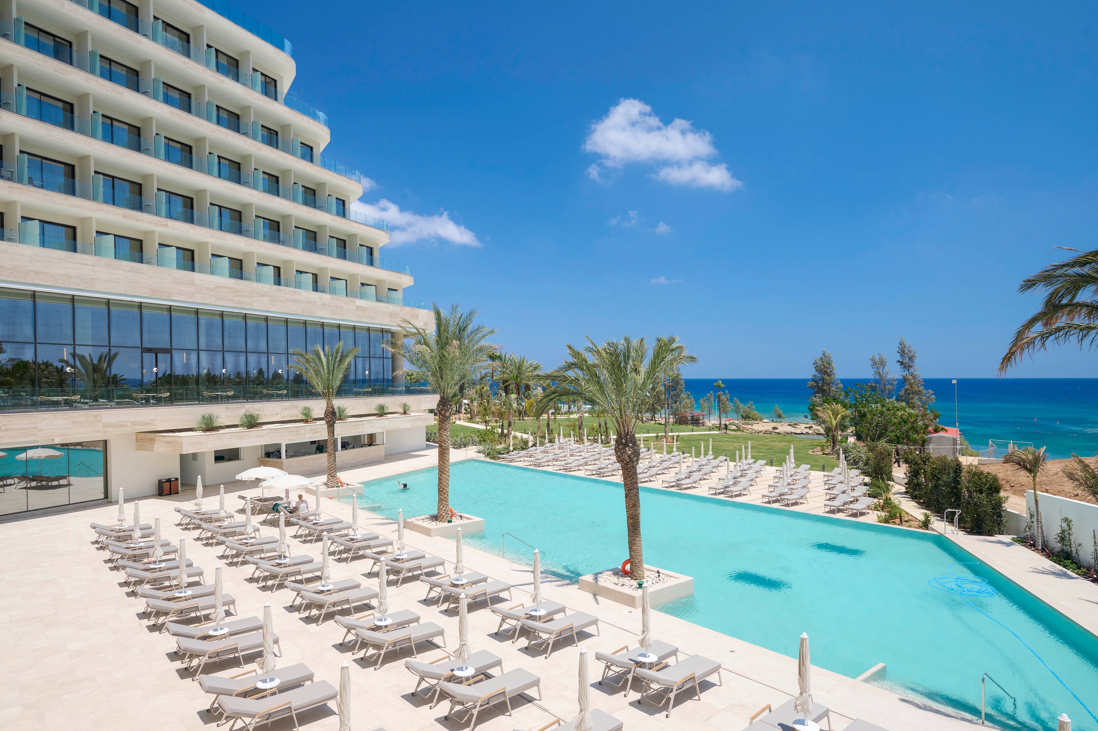 A contemporary multi-story hotel overlooks a large outdoor swimming pool lined with white sun loungers and umbrellas. Tall palm trees are planted around the pool area, adding a tropical feel to the resort. Beyond the hotel grounds, the blue ocean and clear sky create a bright, sunny coastal backdrop. The scene suggests a relaxing vacation destination with ample space for guests to sunbathe and swim.