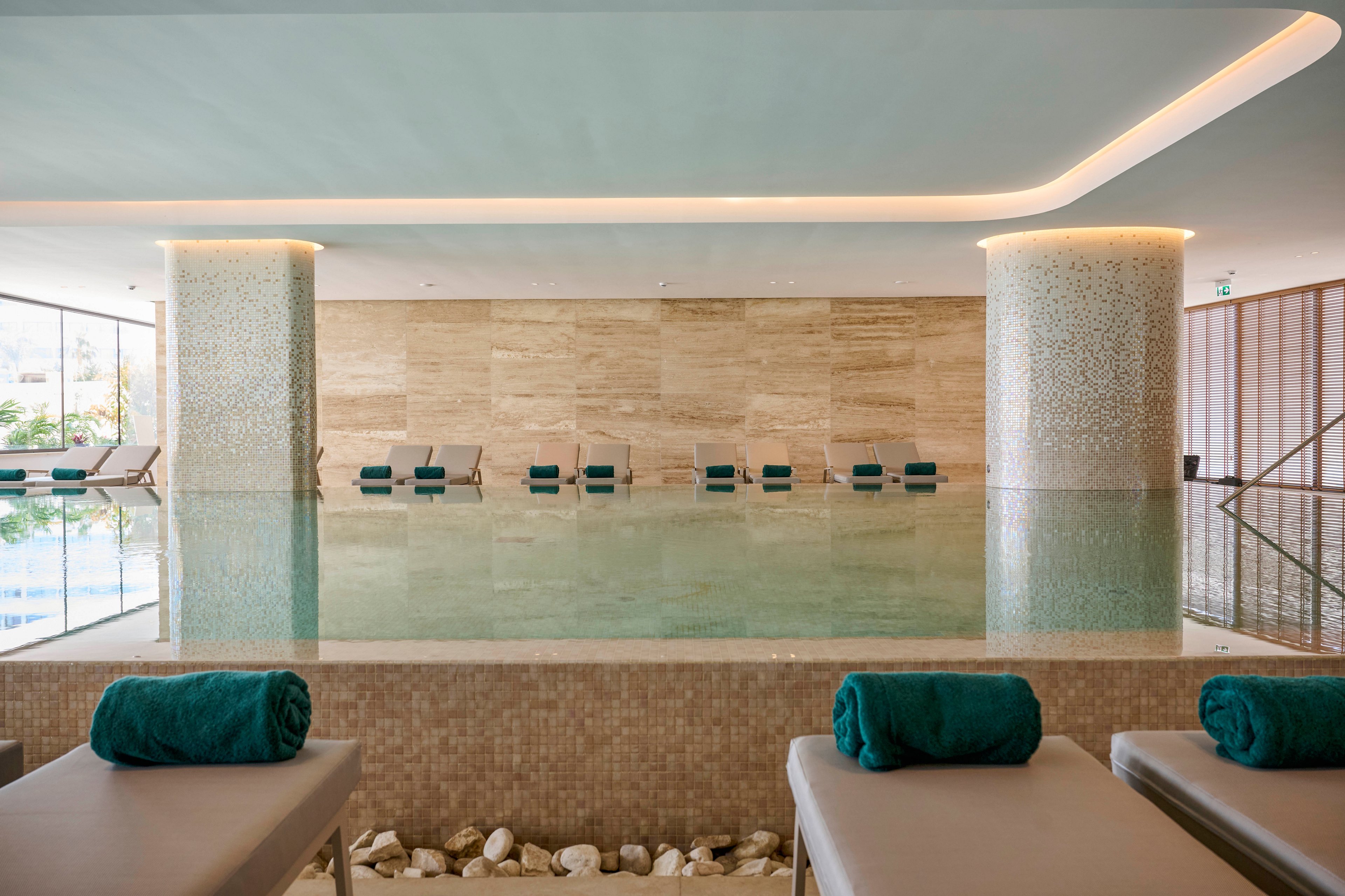 An elegant indoor spa pool is surrounded by cushioned loungers topped with neatly rolled teal towels. The space features smooth stone walls, mosaic-tiled columns, and a calm reflective water surface. Natural light filters in through large windows, enhancing the serene and minimalist design. The overall atmosphere suggests relaxation, wellness, and a high-end resort or hotel setting.