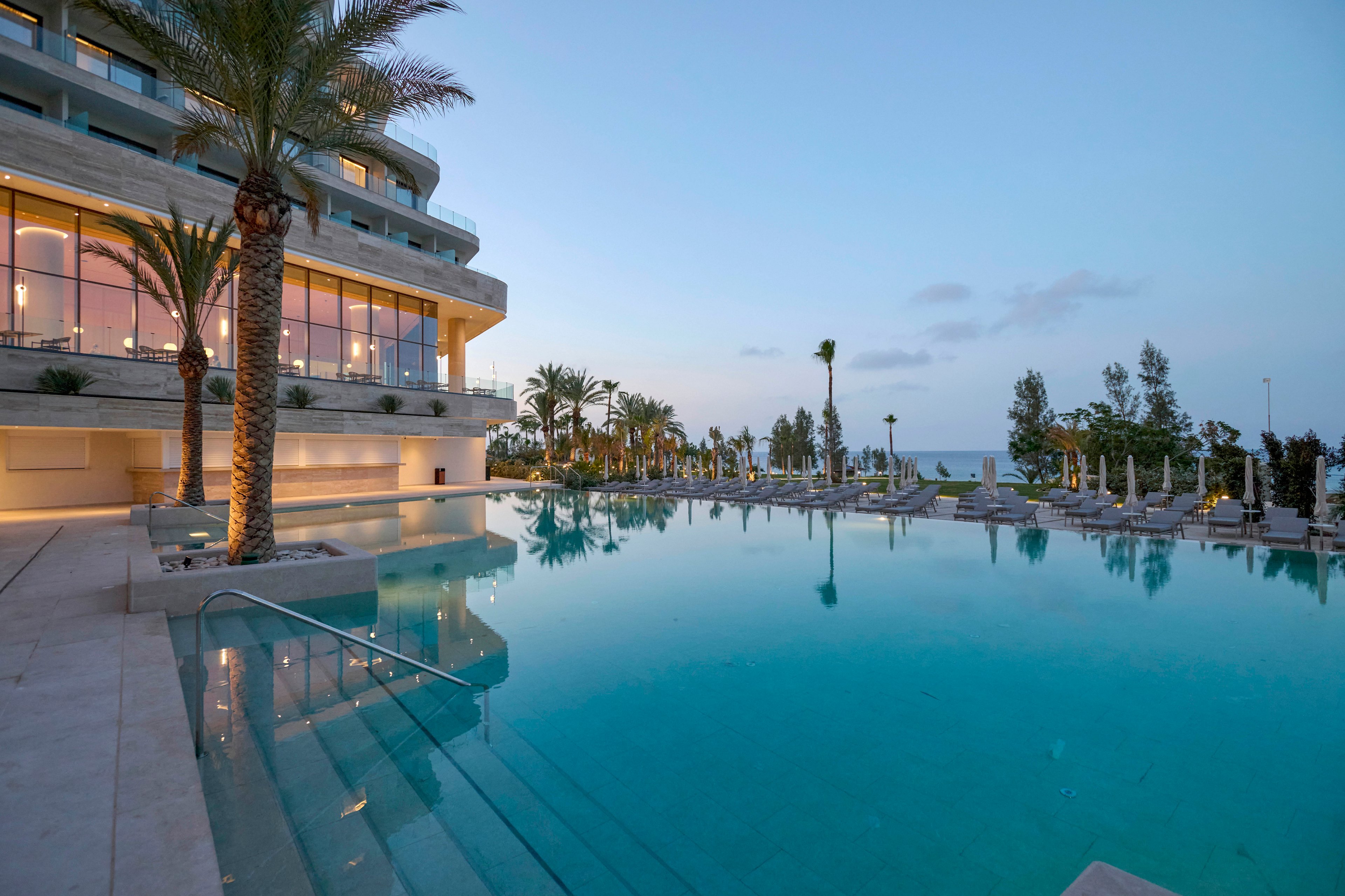 A modern luxury hotel overlooks a large infinity-style swimming pool at dusk. Warm interior lights glow through the building’s glass windows, reflecting softly on the calm water. Palm trees and neatly arranged poolside loungers frame the scene, leading the eye toward the distant sea horizon. The tranquil evening sky and subtle reflections create a serene, upscale resort atmosphere.