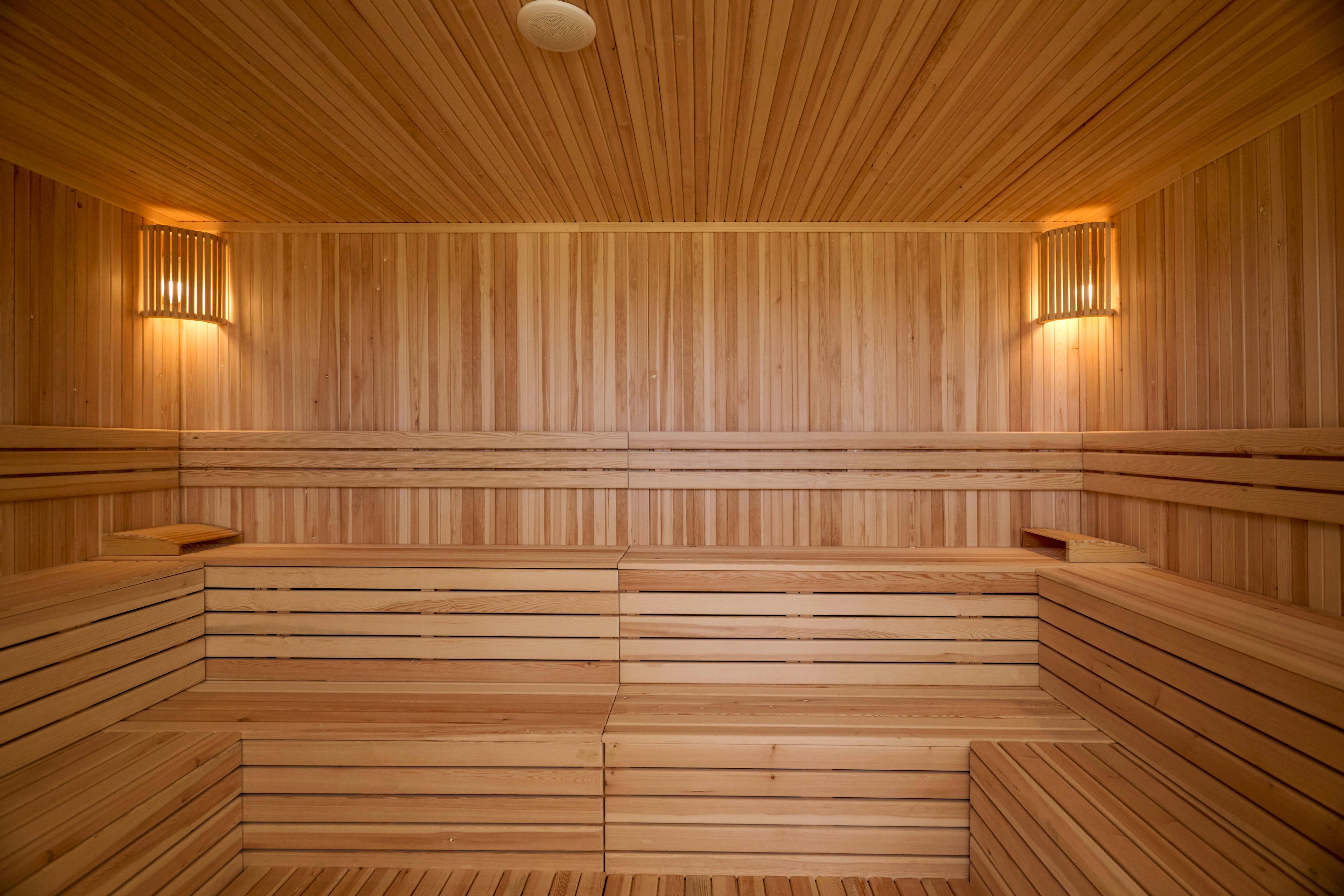 Image of a clean, empty sauna room lined entirely with light wooden panels. Tiered wooden benches run along the walls, creating multiple seating levels. Two warm wall-mounted lights illuminate the space, emphasizing the natural wood grain. The overall atmosphere is calm, minimal, and inviting, suggesting relaxation and wellness.