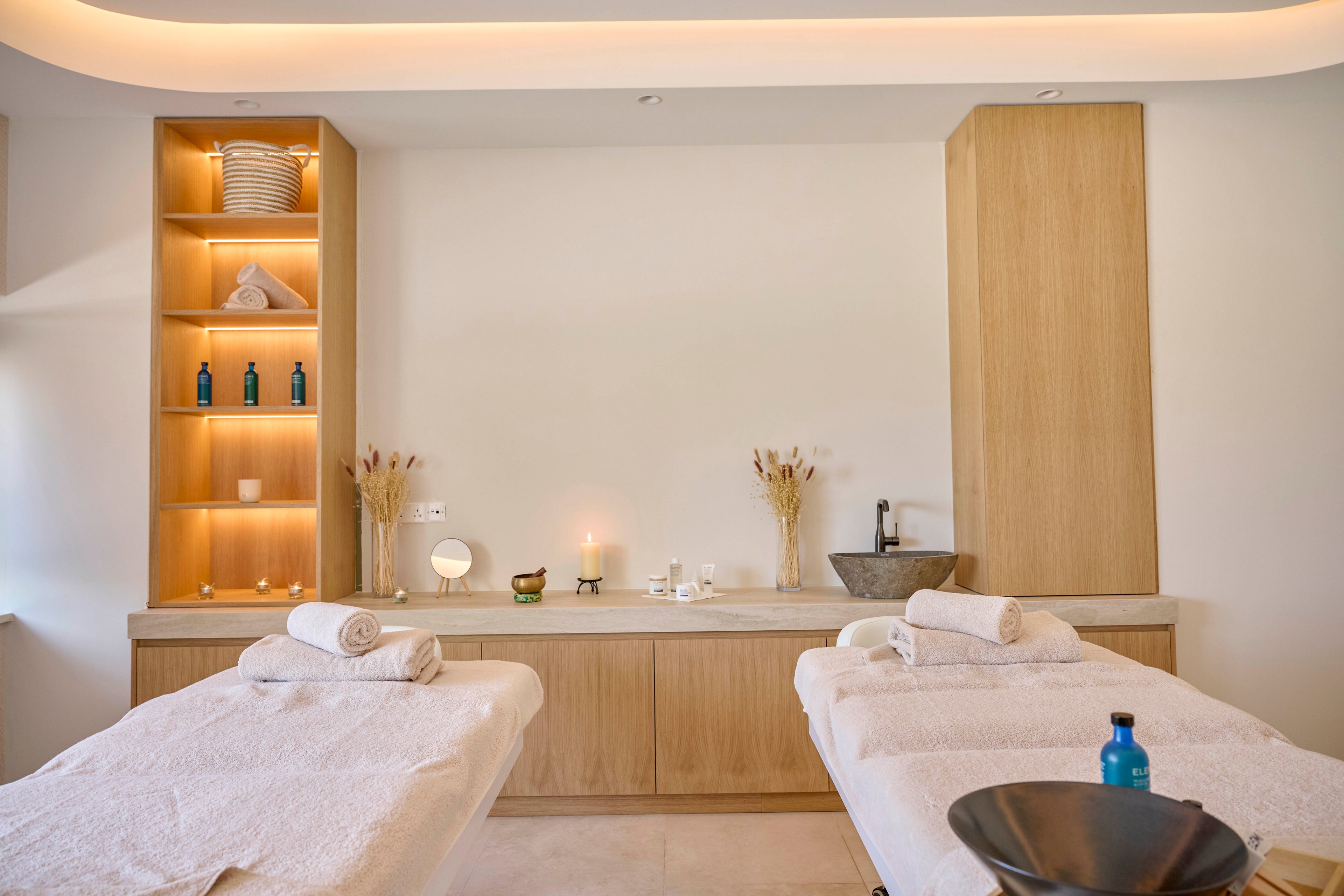 A tranquil spa treatment room features two massage tables neatly prepared with white towels. Built-in wooden cabinetry at the back wall displays bottles, towels, and decorative objects softly lit by warm accent lighting. A candle glows on the counter beside dried floral arrangements and a stone sink, enhancing the relaxing atmosphere. Neutral colors and minimalist decor create a calm, luxurious wellness setting.