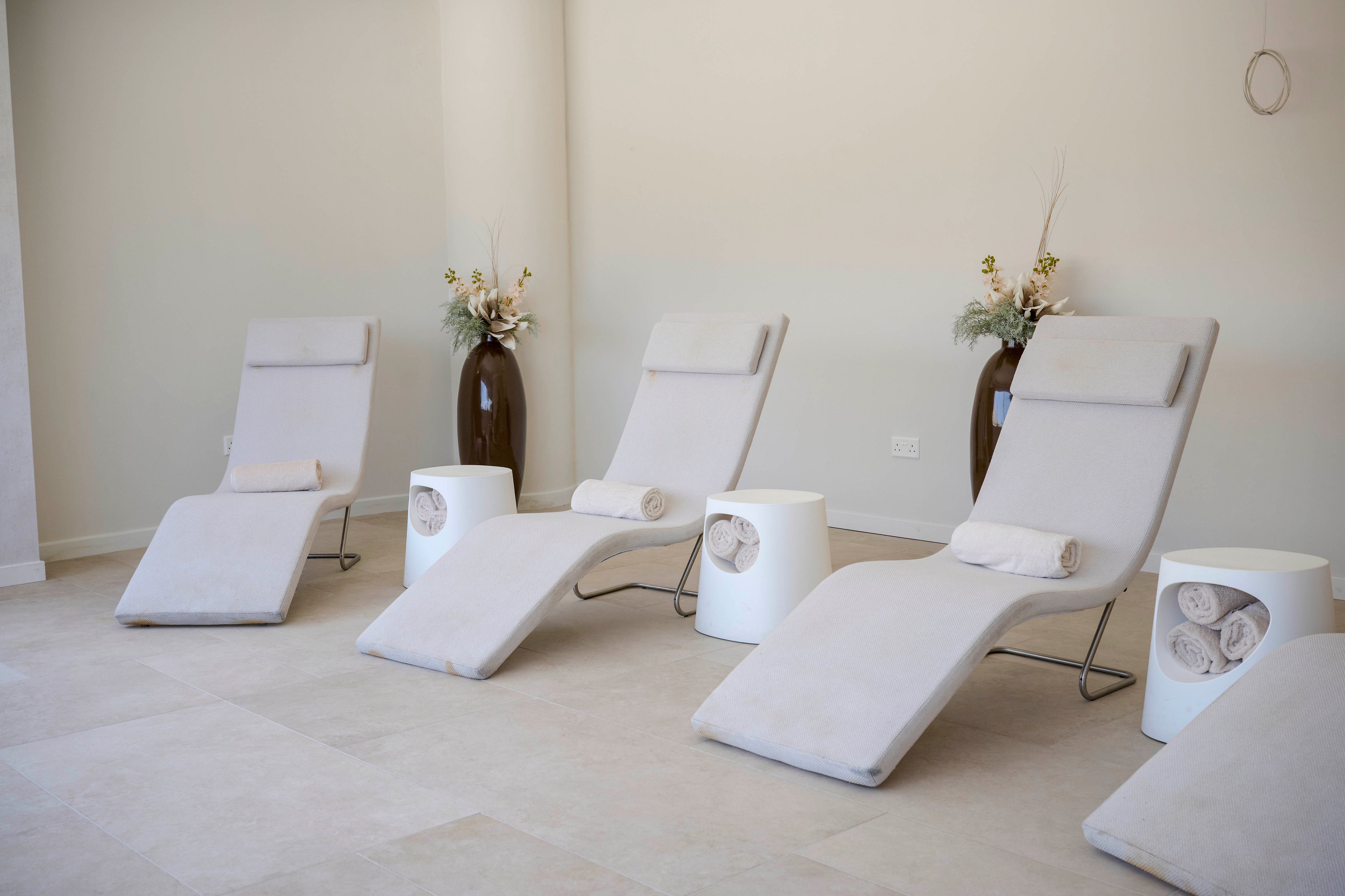 A row of modern, cushioned loungers is arranged in a minimalist spa relaxation room. Each chair is paired with a small white side table holding neatly rolled towels. Neutral beige and white tones dominate the space, creating a calm and serene atmosphere. Tall vases with simple floral arrangements add a subtle decorative touch against the plain walls.