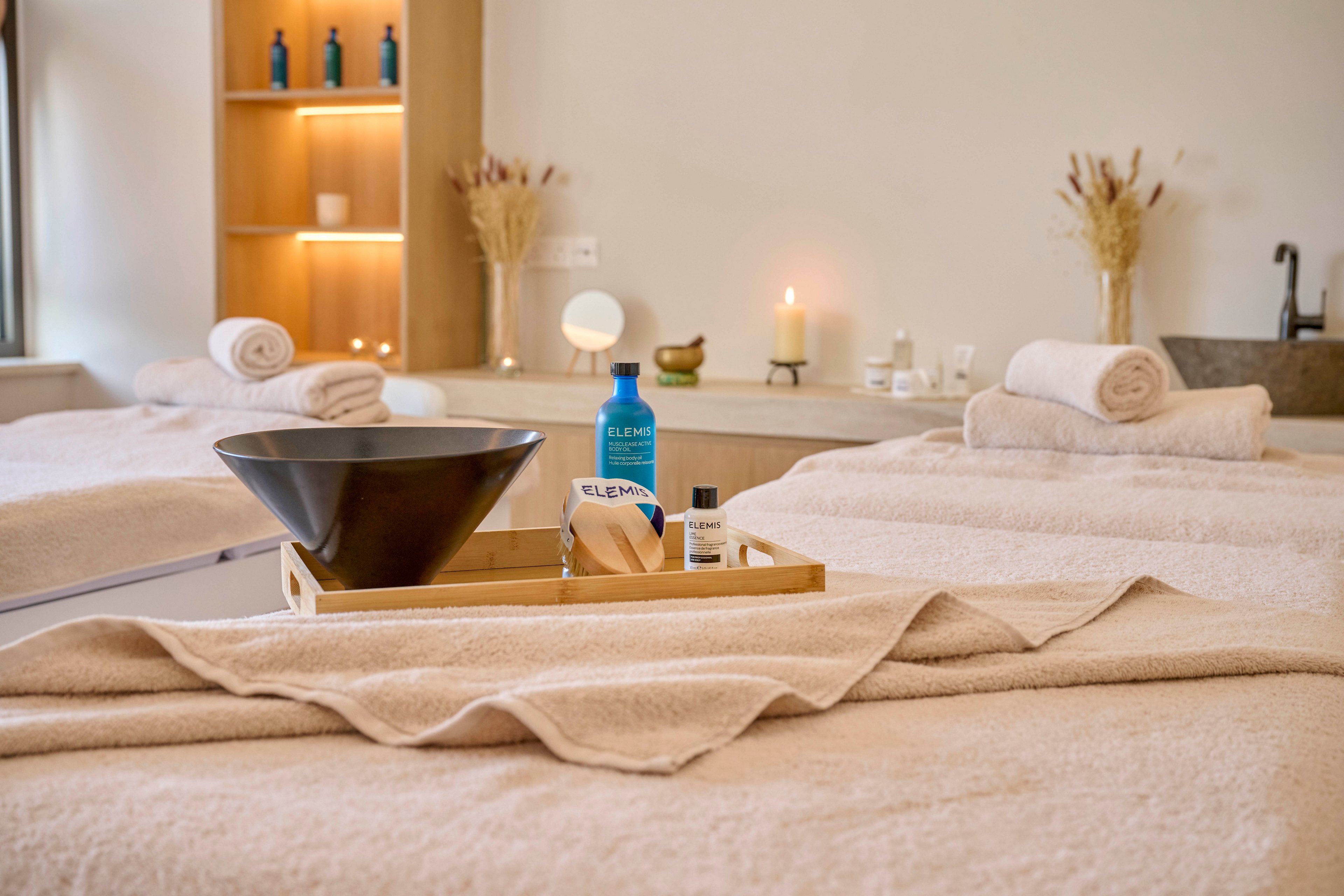 A serene spa treatment room features neatly arranged massage beds covered in soft beige towels. In the foreground, a wooden tray holds a black bowl, a blue bottle, a small white bottle, and a natural sponge. Rolled towels, dried floral arrangements, and softly glowing candles create a calming atmosphere in the background. Shelving with additional bottles and decor enhances the relaxing, minimalist interior design.