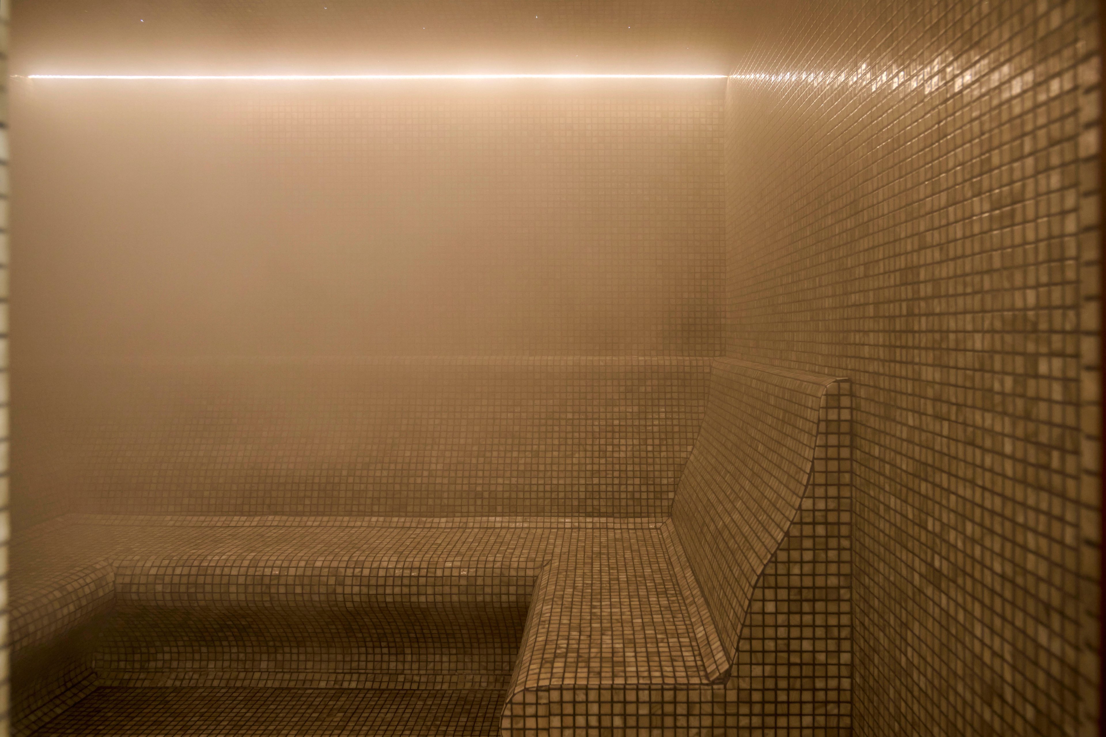 A tiled steam room interior is filled with dense mist, partially obscuring the walls and bench. The space features small square tiles in neutral tones covering the seating and surrounding surfaces. A linear light strip runs along the upper edge of the wall, softly illuminating the steam. The overall atmosphere suggests relaxation, wellness, and spa-like tranquility.