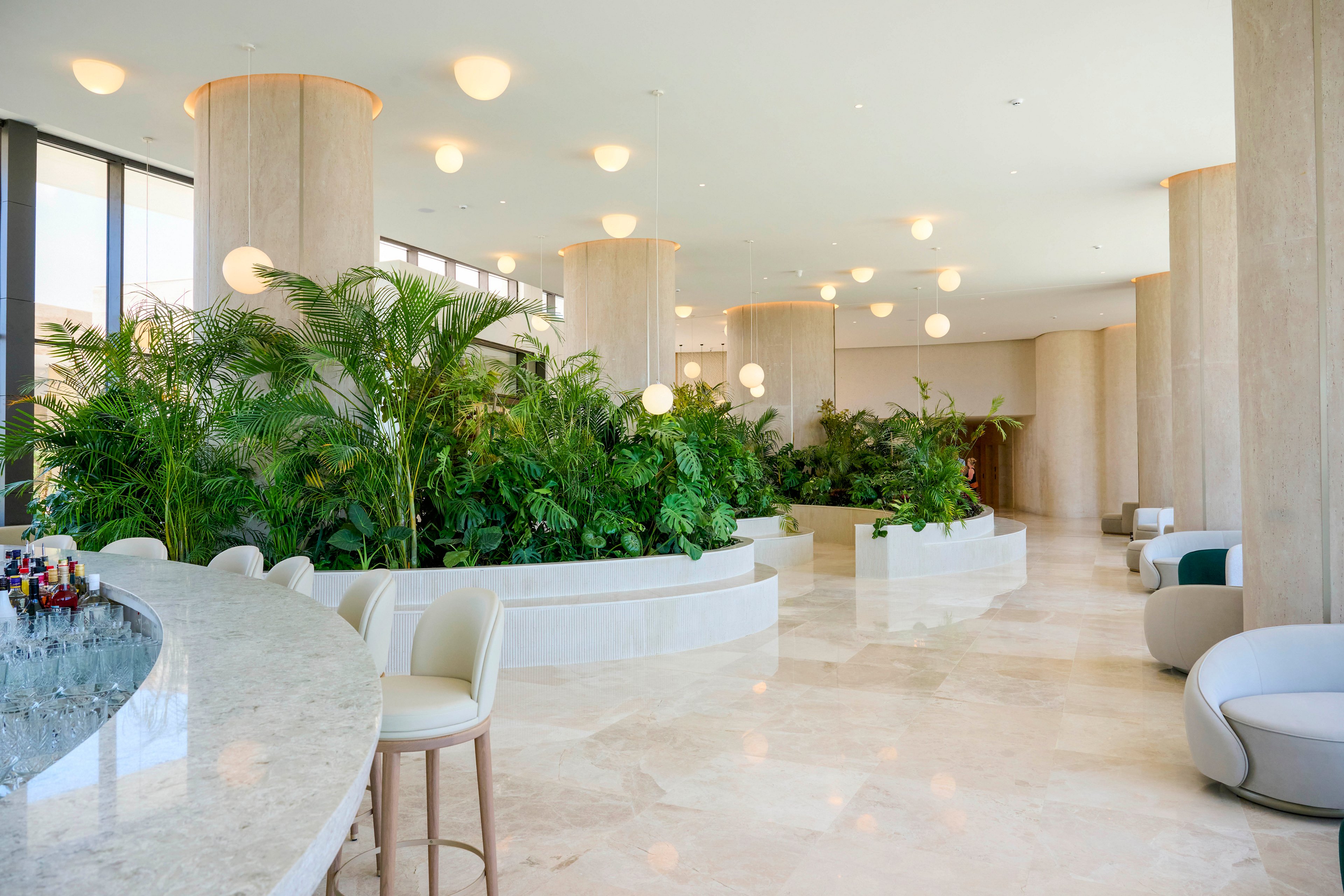 A spacious modern lobby features a curved marble bar and plush seating. Large planters filled with lush green plants divide the open area and soften the architecture. Tall columns, polished stone floors, and round ceiling lights create a bright, airy atmosphere. Floor-to-ceiling windows on the side bring in natural light, enhancing the clean, contemporary design.