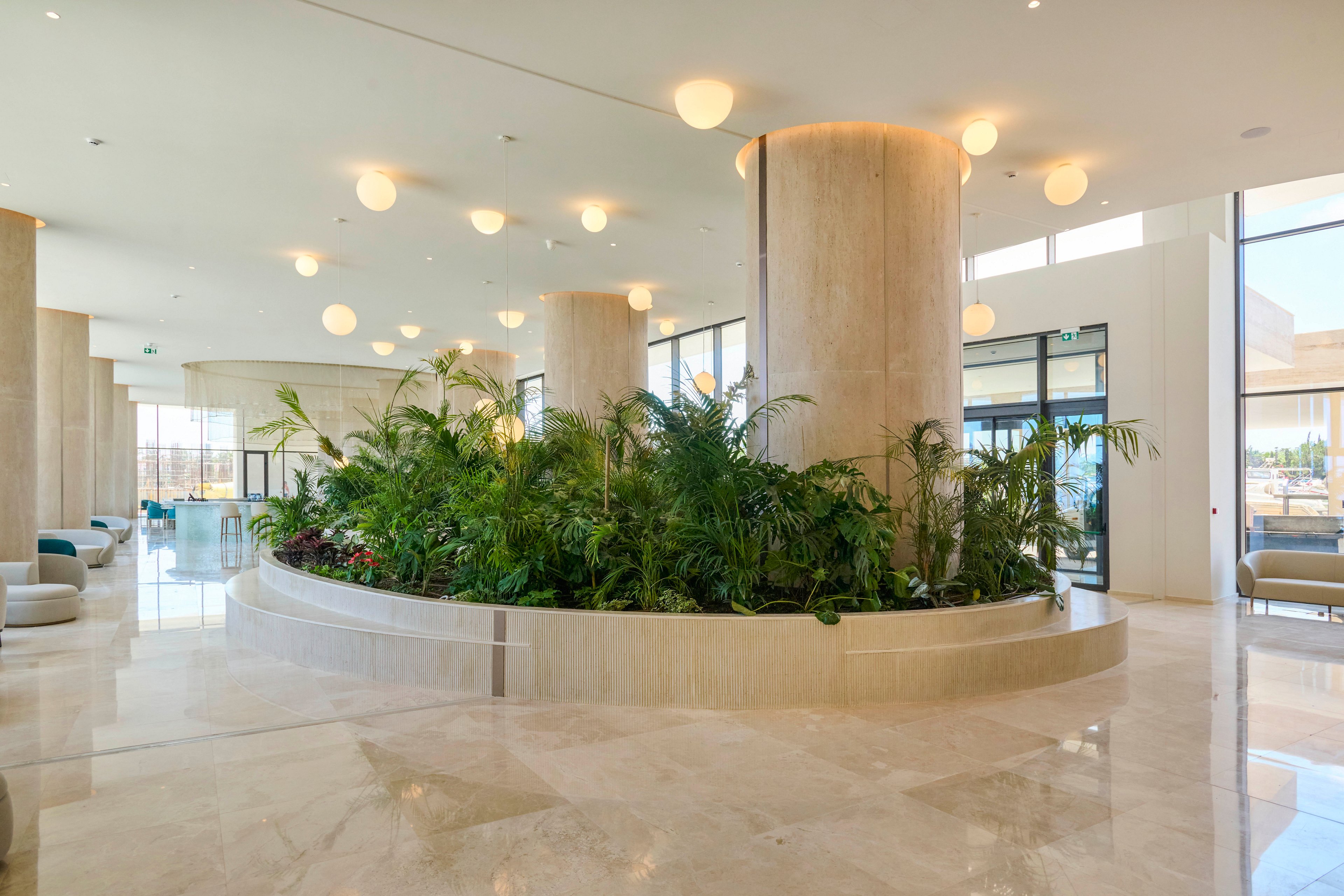 Spacious contemporary lobby interior features a large circular planter filled with lush green tropical plants. Tall cylindrical columns rise through the planter, supporting a high ceiling dotted with round pendant lights. Polished marble or stone flooring reflects the warm ambient lighting, enhancing the airy and luxurious feel. Floor-to-ceiling windows in the background allow natural light to flood the space and offer views of the outdoors.
