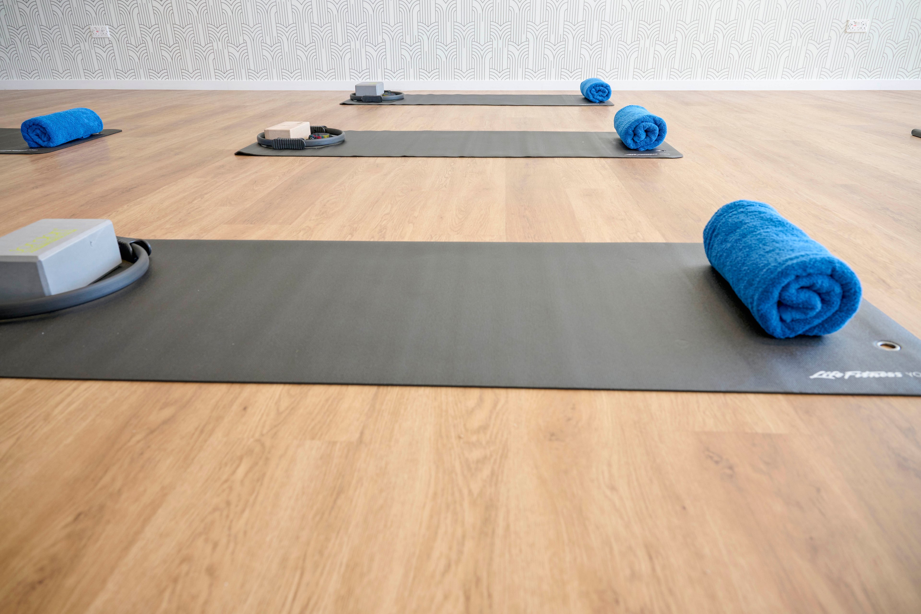 A bright yoga studio features several gray mats laid out in rows on a light wooden floor. Each mat is prepared with a rolled blue towel and small exercise props, suggesting readiness for a class. The room appears clean, minimal, and organized, with soft natural light enhancing the calm atmosphere. No people are present, emphasizing the tranquil, pre-class setting.