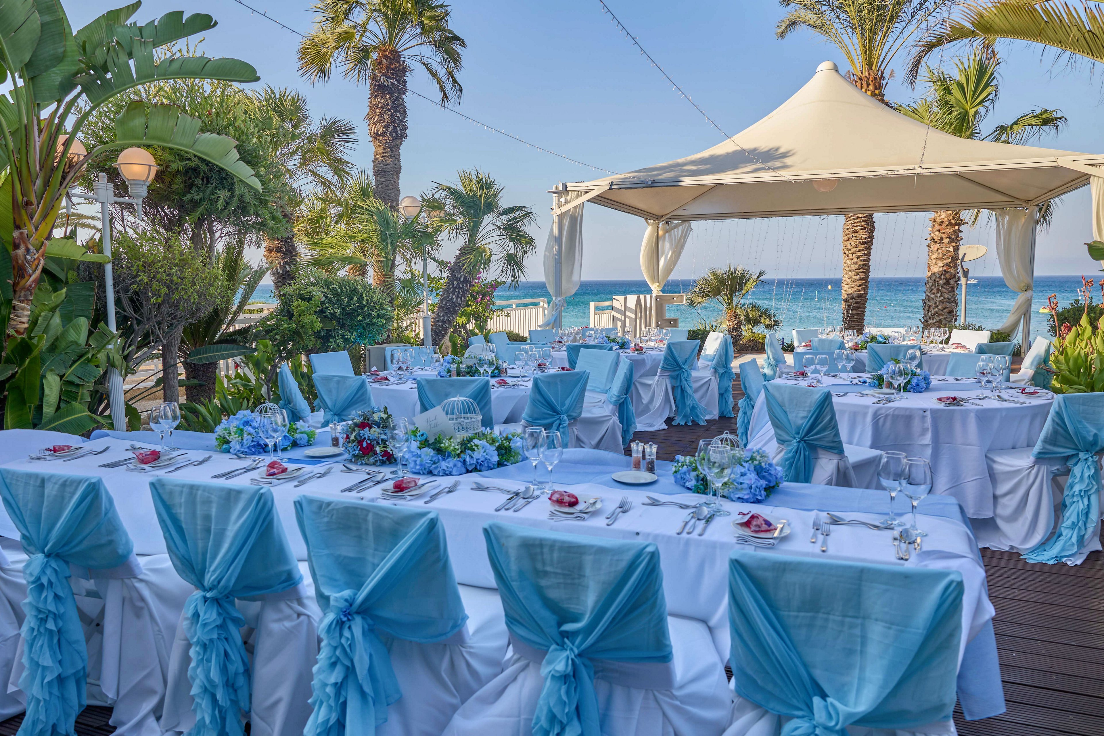 An outdoor wedding reception is set up on a wooden deck overlooking the sea. Round and rectangular tables are covered with white linens and decorated with blue chair sashes and floral centerpieces. A large canopy tent provides shade, while palm trees and lush greenery frame the view of the ocean. The scene suggests a formal celebration in a bright, coastal setting.