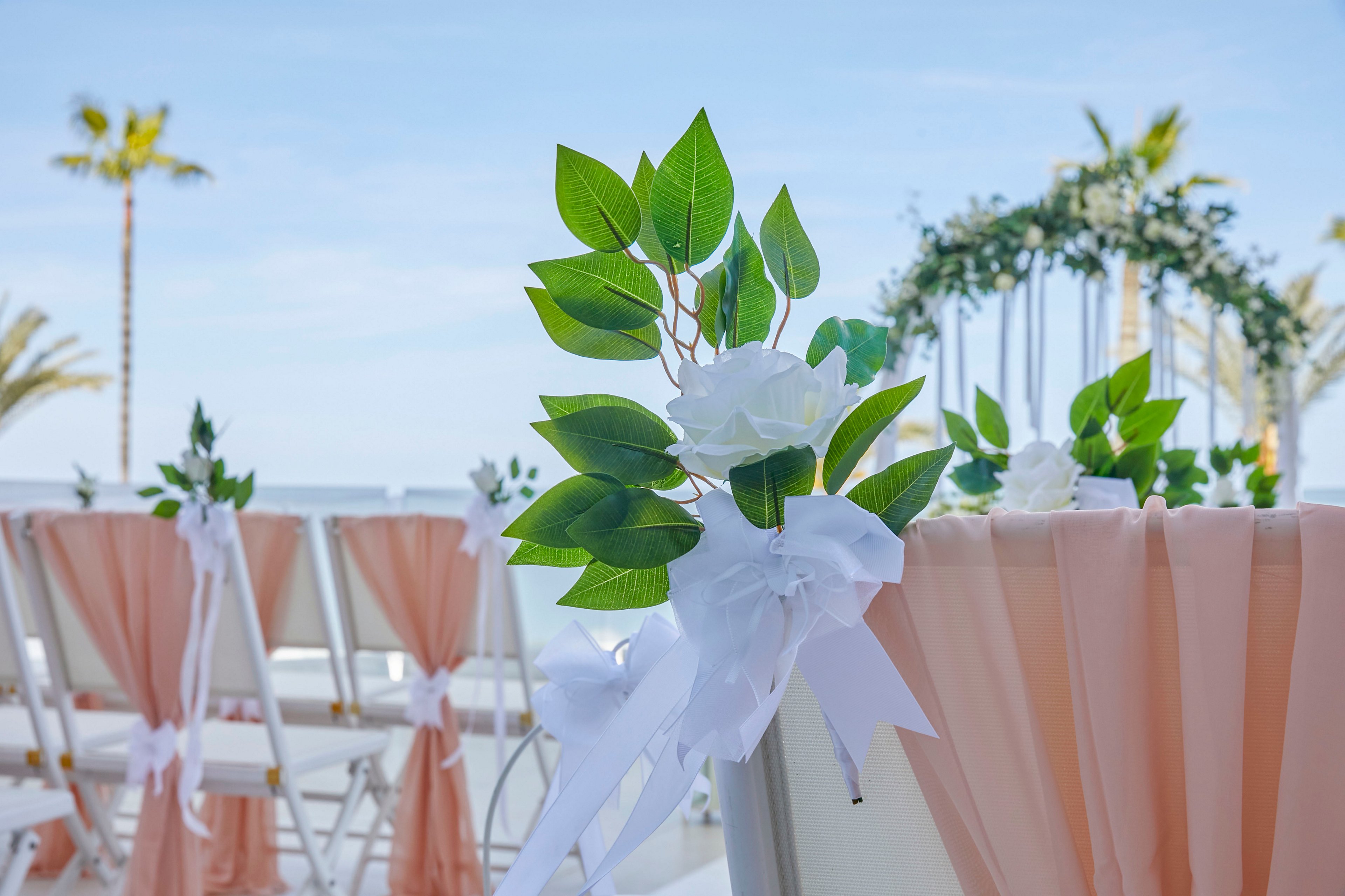 White floral arrangements with green leaves adorn chairs draped in soft peach fabric at an outdoor wedding ceremony. The setting overlooks the ocean under a clear blue sky, with palm trees visible in the background. A floral archway is positioned at the end of the aisle, creating a romantic focal point. The overall scene feels airy, elegant, and serene.