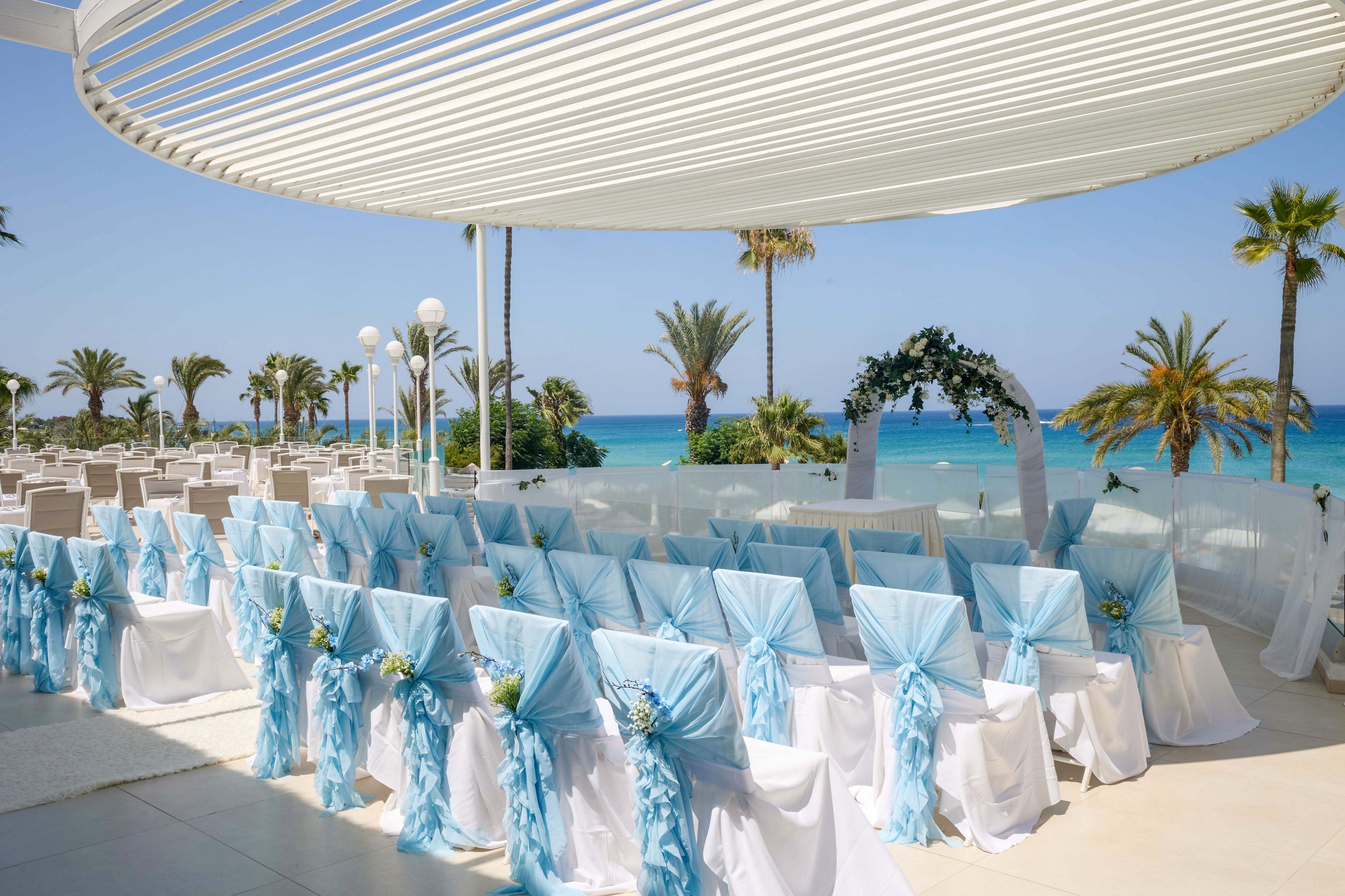 Rows of white chairs with light blue sashes are arranged for a wedding ceremony overlooking a bright blue sea. A floral arch stands at the front, framed by palm trees and a clear sky. The venue is shaded by a modern white canopy, creating a fresh and airy atmosphere. Decorative flowers and soft pastel colors enhance the romantic coastal setting.