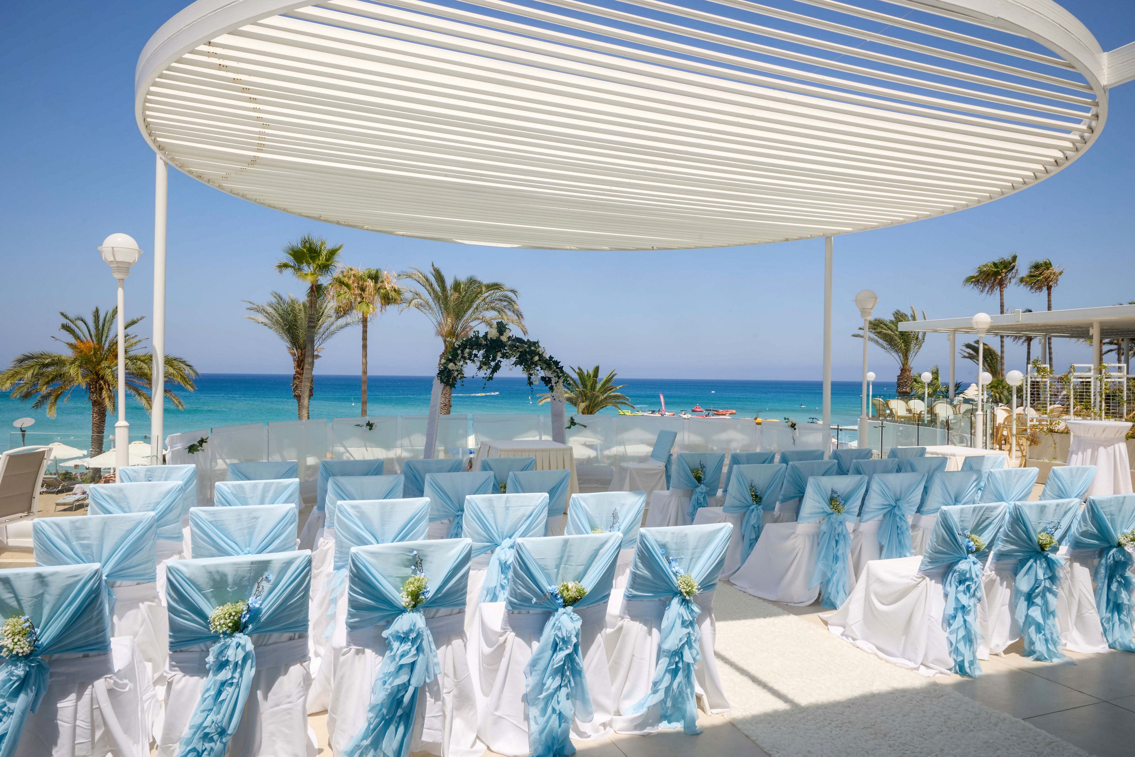 Rows of white chairs with light blue sashes are arranged for a wedding ceremony under a modern pergola. The setting overlooks a bright turquoise sea with palm trees and a clear blue sky in the background. Decorative flowers are attached to the backs of the chairs, enhancing the festive atmosphere. The scene suggests a sunny, elegant outdoor event at a coastal venue.