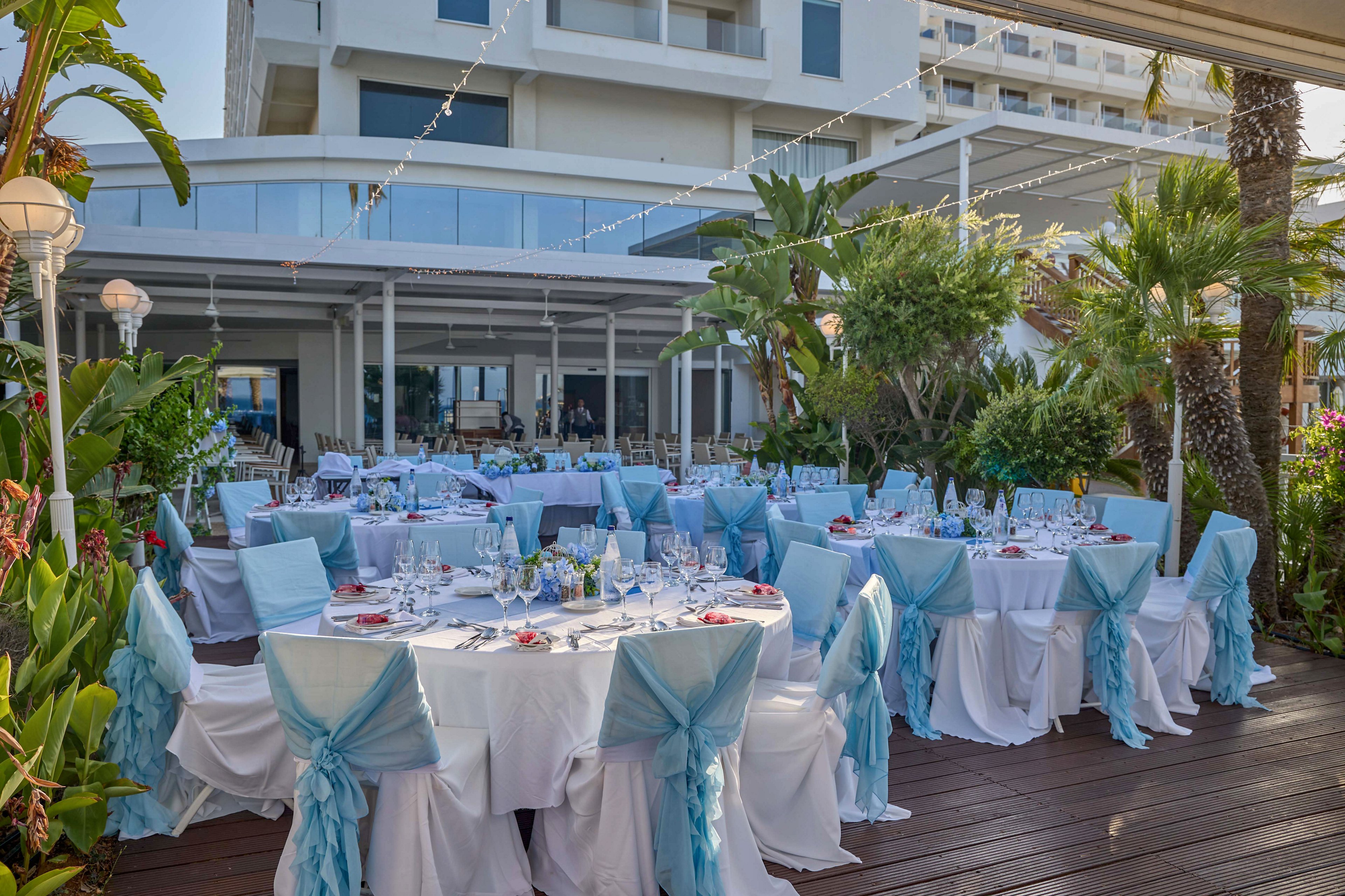 Elegant round tables are arranged for an outdoor reception with white linens and light blue chair sashes. The setting appears to be a terrace beside a modern hotel or resort, surrounded by palm trees and lush greenery. Each table is set with glassware, plates, and small red accents, suggesting a formal event such as a wedding or celebration. String lights overhead add a festive touch to the bright, coastal atmosphere.