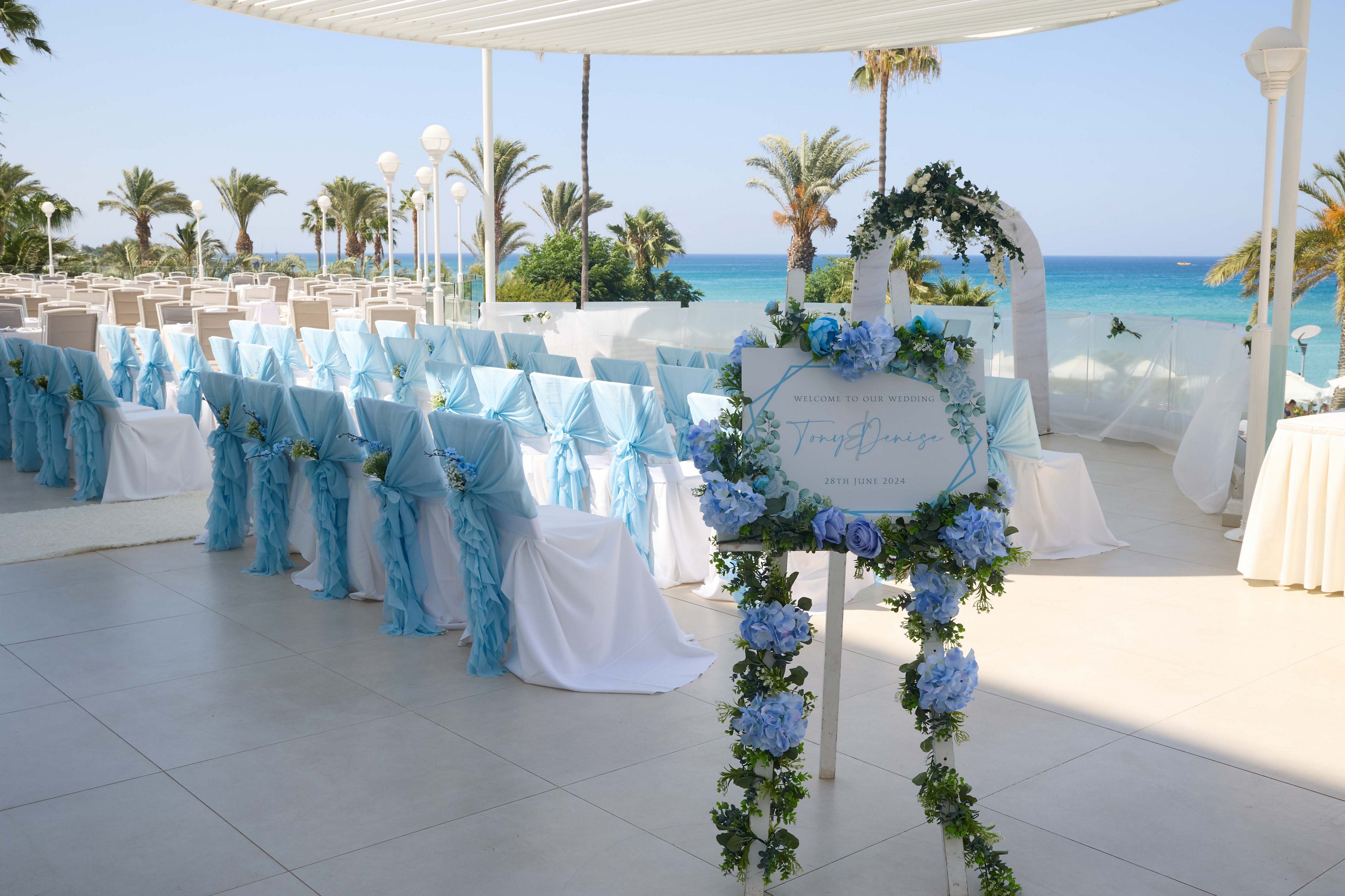 Rows of chairs draped in light blue fabric are arranged for a wedding ceremony overlooking a turquoise sea. A floral wreath with blue and white flowers frames a decorative sign at the entrance to the aisle. Palm trees and white columns line the open-air terrace, creating a bright coastal atmosphere. The scene is bathed in natural sunlight, emphasizing the romantic and serene beachside setting.