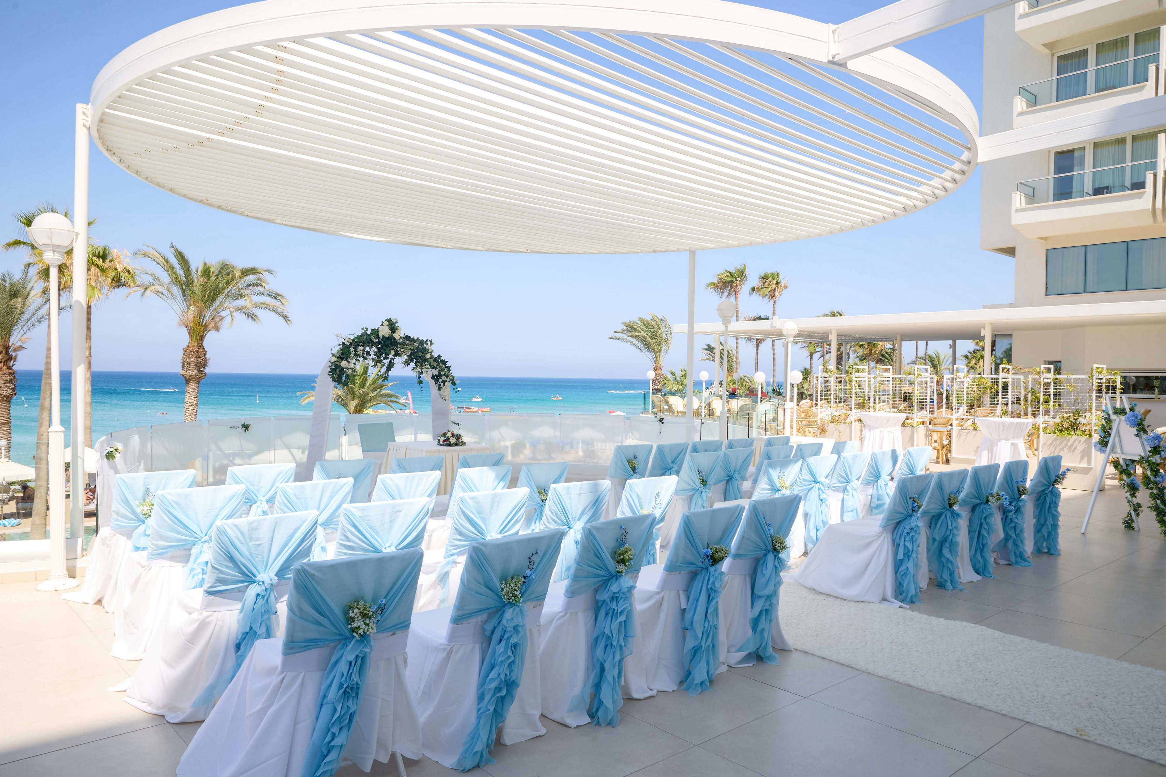 Rows of white chairs with light blue sashes are arranged for a wedding ceremony on a terrace overlooking the ocean. A modern white pergola shades the seating area, while palm trees and a clear blue sky frame the coastal backdrop. Floral decorations accent the aisle and arch, adding a romantic touch to the bright, airy setting. The scene conveys a serene, festive atmosphere ideal for a beachside celebration.