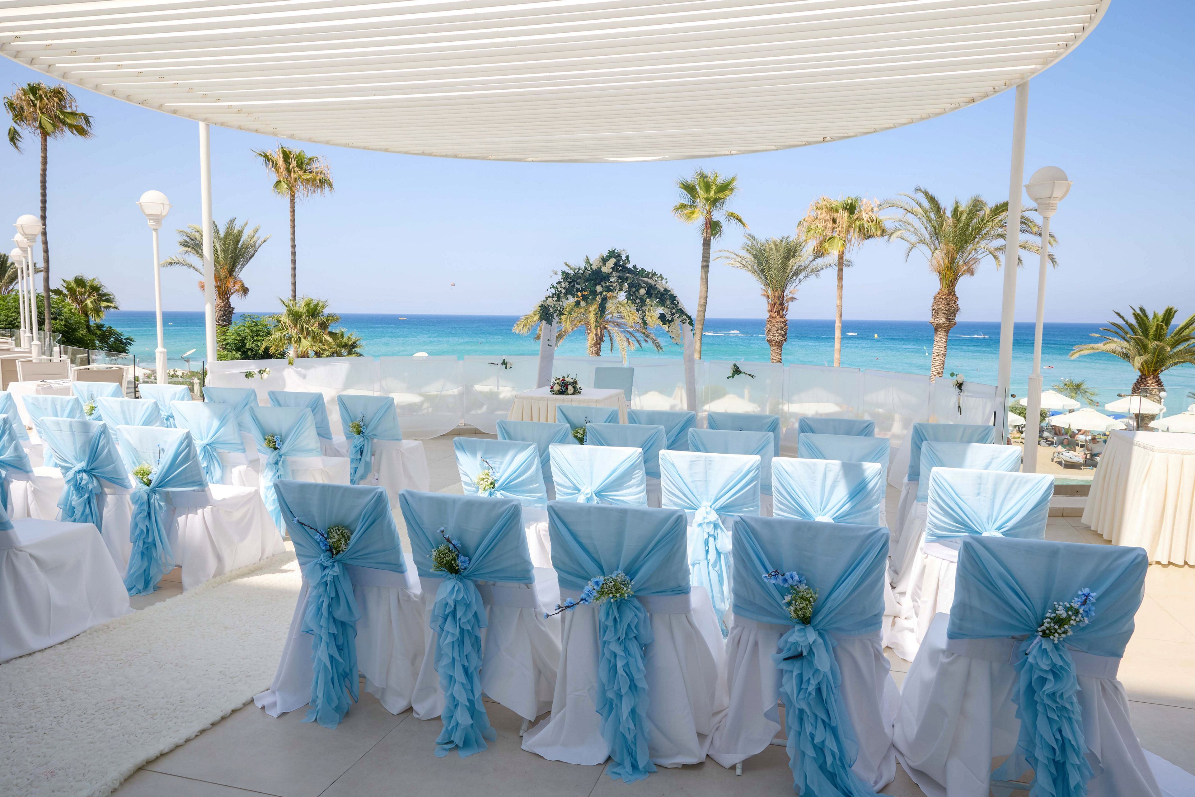 Rows of white chairs decorated with light blue fabric are arranged for a wedding ceremony overlooking a tropical beach. The seating area is set under a modern white canopy with a clear view of turquoise water and palm trees. Small floral arrangements accent the chair backs, adding a delicate touch to the decor. The bright daylight and soft pastel colors create a romantic and serene atmosphere.