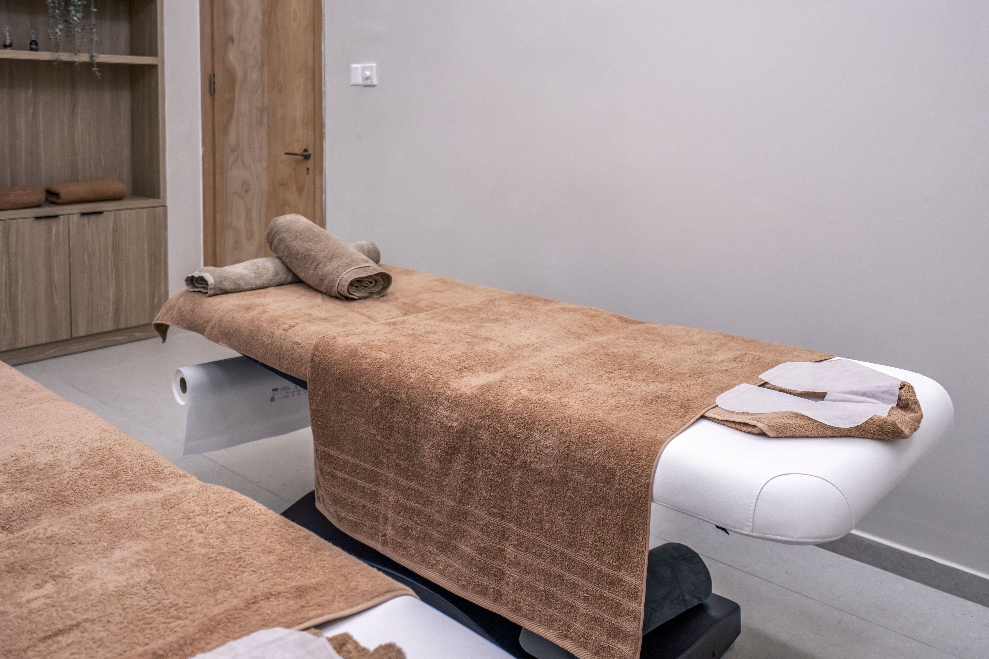 A spa treatment room features two massage tables covered with soft brown towels. Rolled towels are neatly placed at the head of each table, suggesting preparation for a massage session. The neutral walls and wooden cabinetry create a calm, minimalist environment. The overall setting conveys cleanliness, relaxation, and professional spa care.