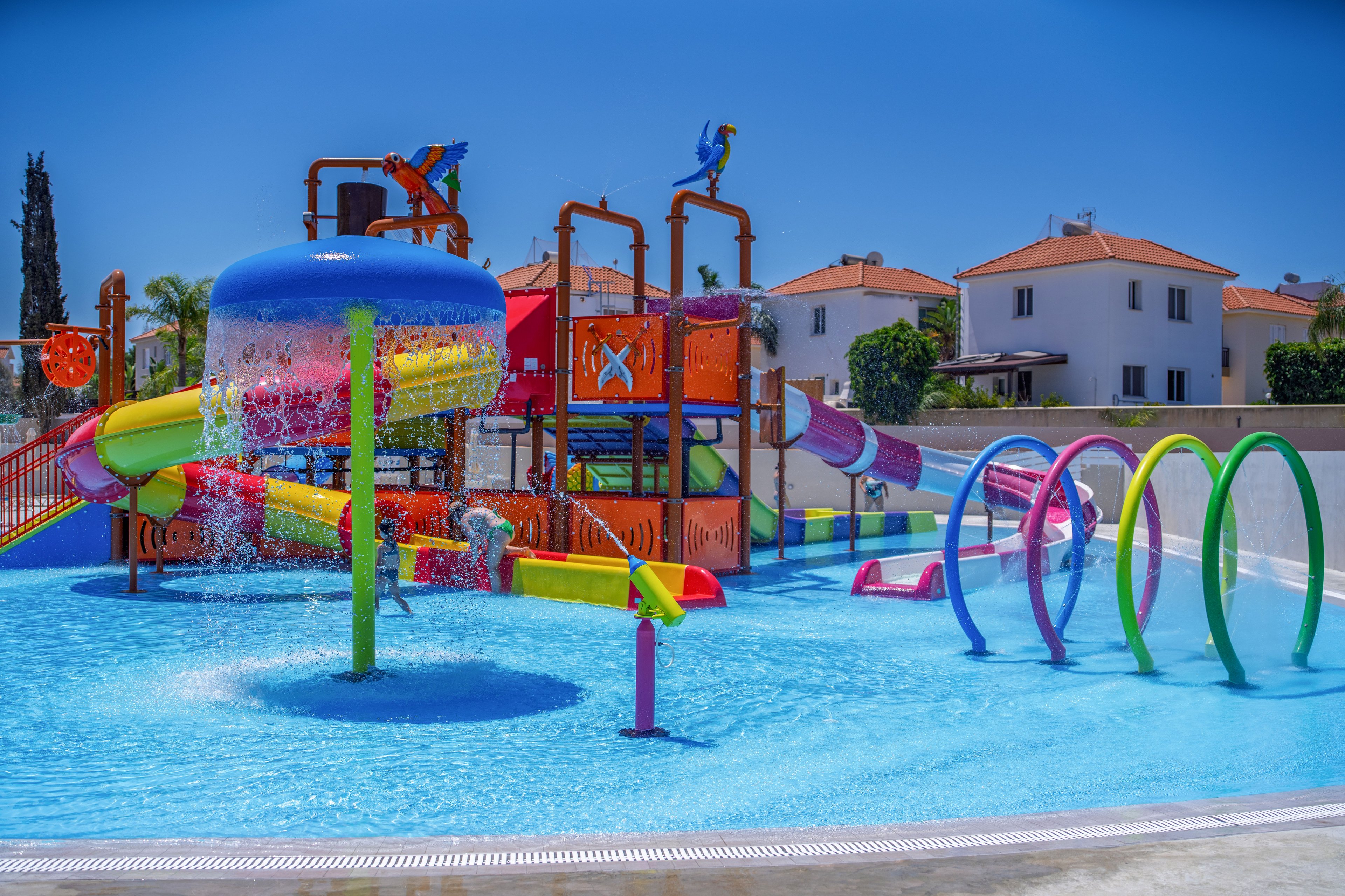 A vibrant children’s water playground features colorful slides, fountains, and climbing structures in a shallow pool. Water sprays from multiple pipes and arches, creating a playful and refreshing scene under a clear blue sky. In the background, white houses with red roofs line the edge of the park, emphasizing a suburban resort setting. The bright colors and sparkling water convey a fun, family-friendly atmosphere.