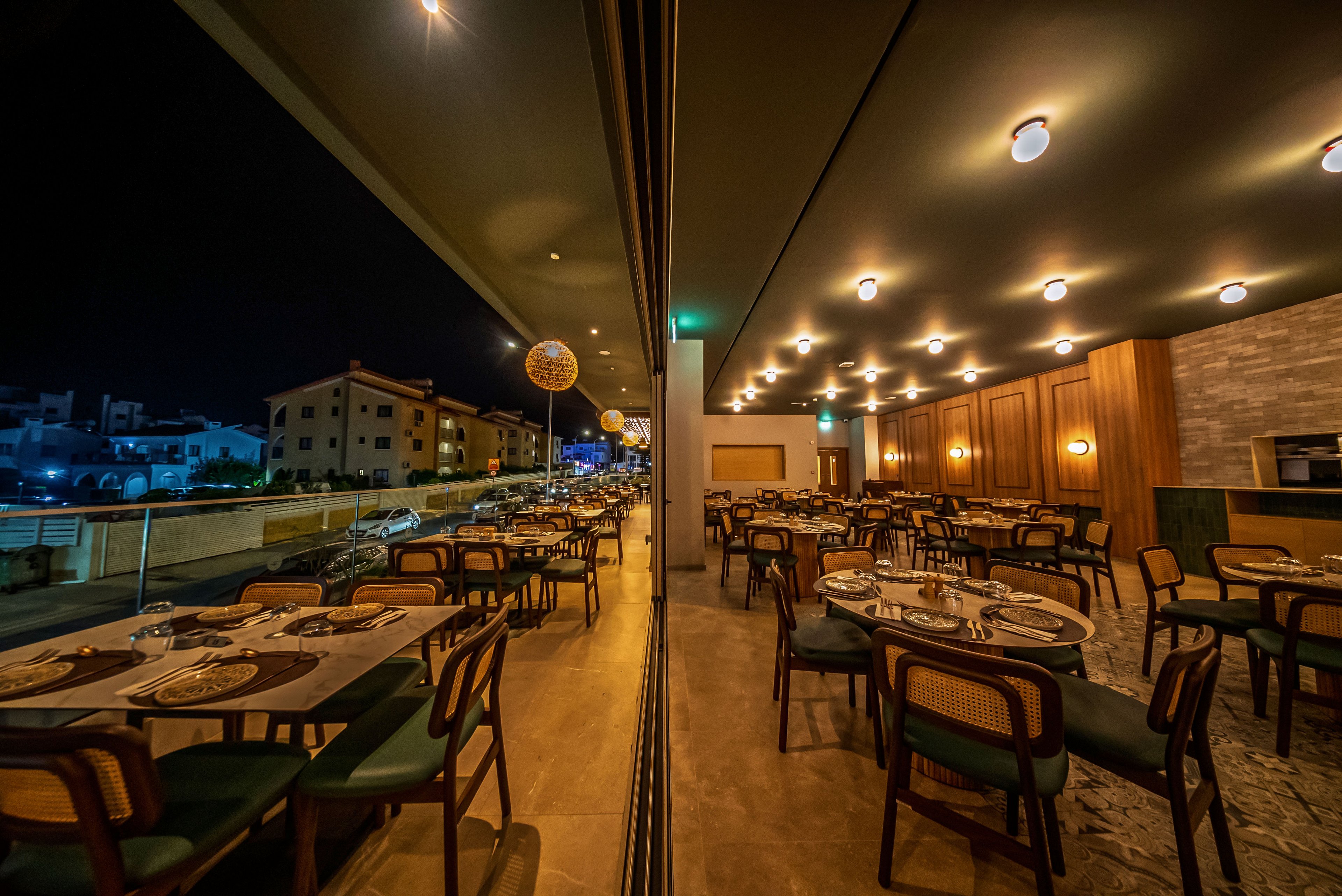 A contemporary restaurant dining area is shown at night with neatly arranged tables and chairs. A large glass wall creates a strong reflection, visually doubling the interior space. Warm ceiling lights and wooden wall panels give the room a cozy, inviting atmosphere. Outside, a nighttime cityscape with residential buildings is visible along the street.