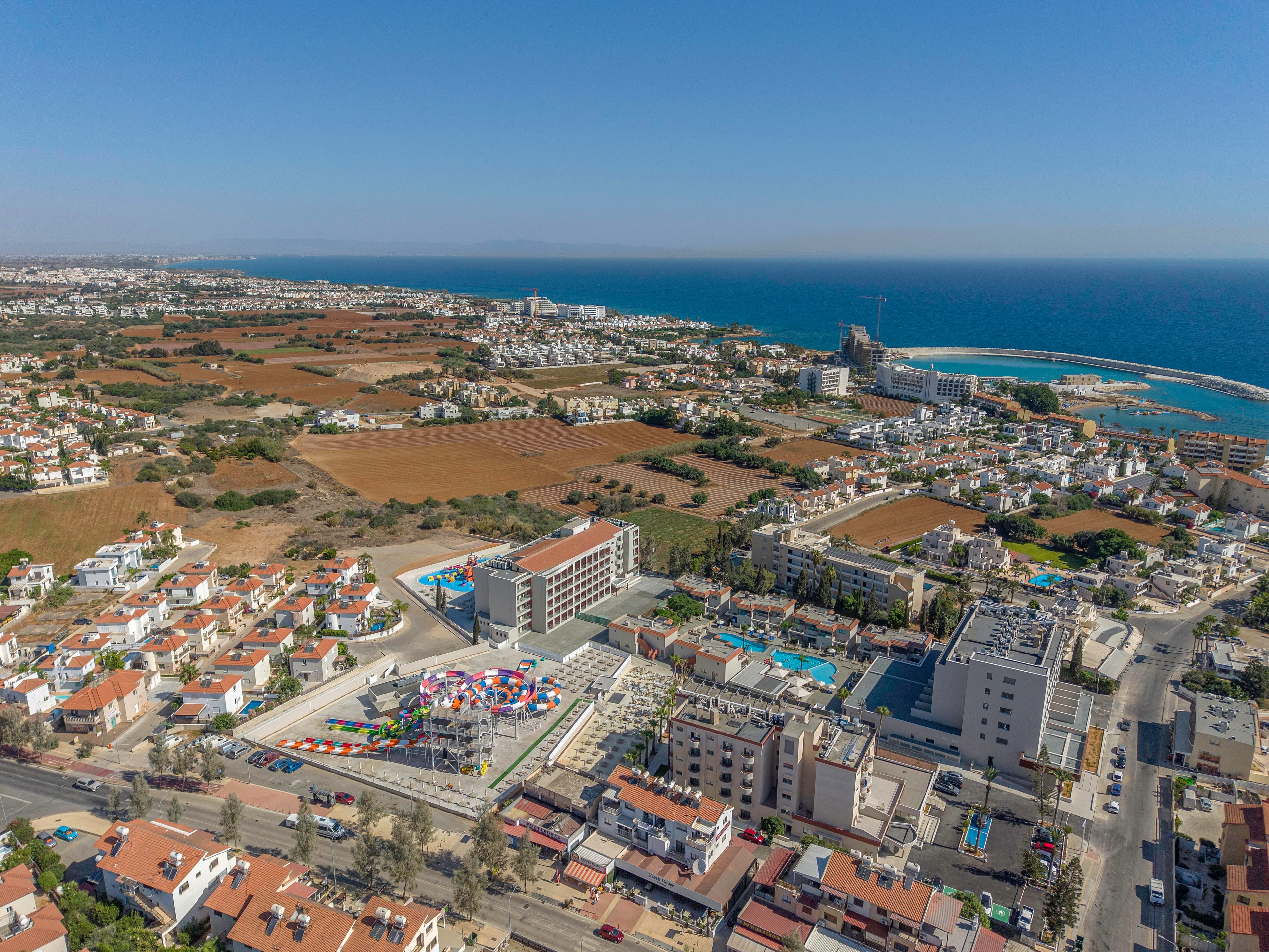 A coastal town is captured from an elevated aerial perspective, showing a mix of residential buildings and open fields. In the foreground, a colorful playground or amusement area stands out among mid-rise apartment blocks. The coastline with a curved harbor and blue sea stretches along the right side of the image. Clear skies and bright sunlight highlight the orderly streets and the contrast between urban development and surrounding farmland.