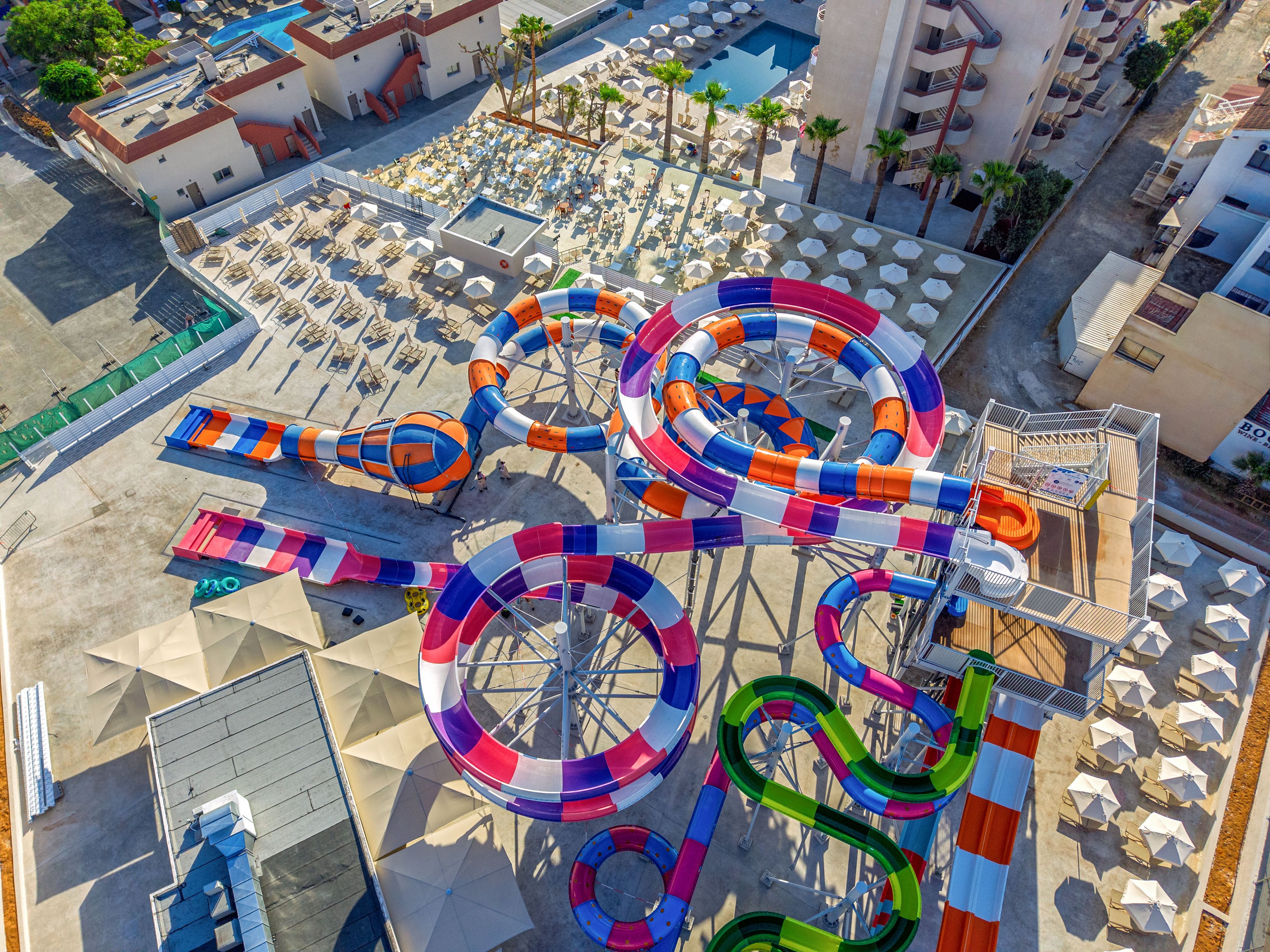 A vibrant rooftop water park features multiple intertwined water slides in bright red, orange, purple, and green colors. The slides twist and loop above a resort area filled with neatly arranged lounge chairs and umbrellas. Surrounding buildings and palm trees frame the aquatic attraction, suggesting a sunny vacation destination. The aerial perspective highlights the complex layout of the slides and the leisure spaces below.